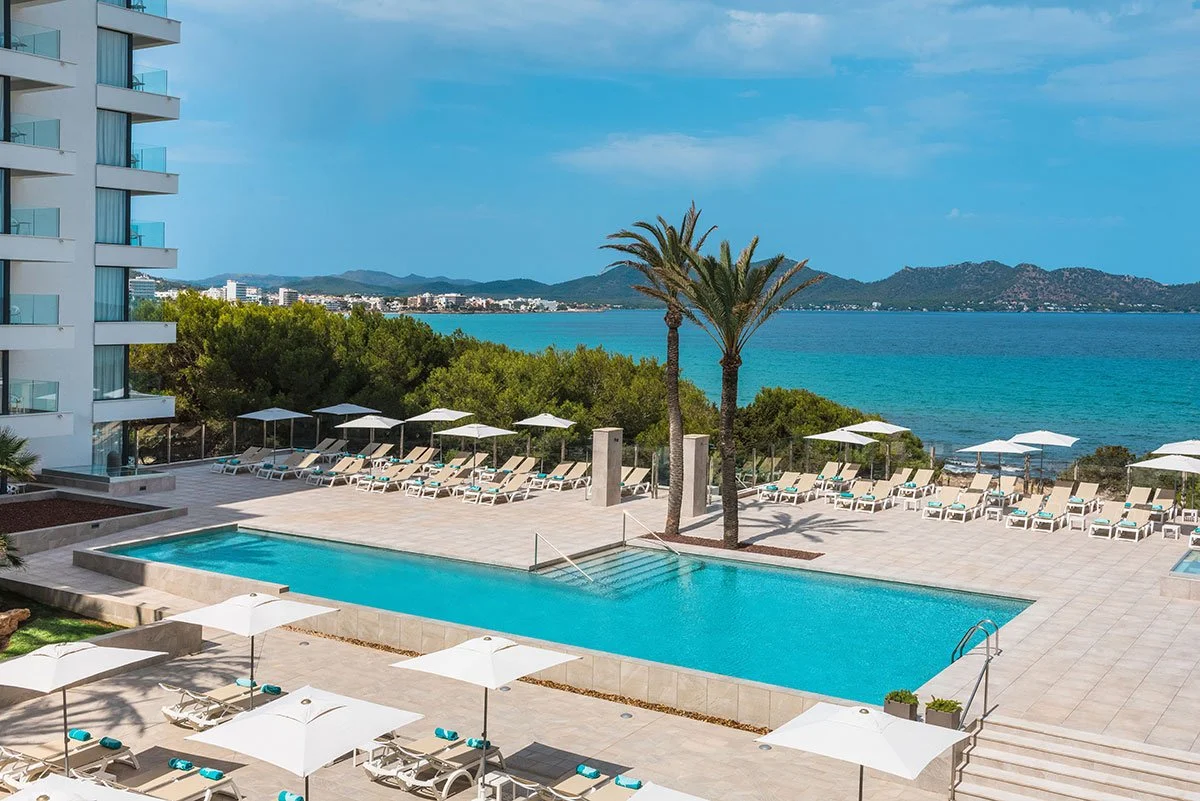 Pool area with lounge chairs and umbrellas overlooking the ocean, with palm trees, a building, and distant mountains.