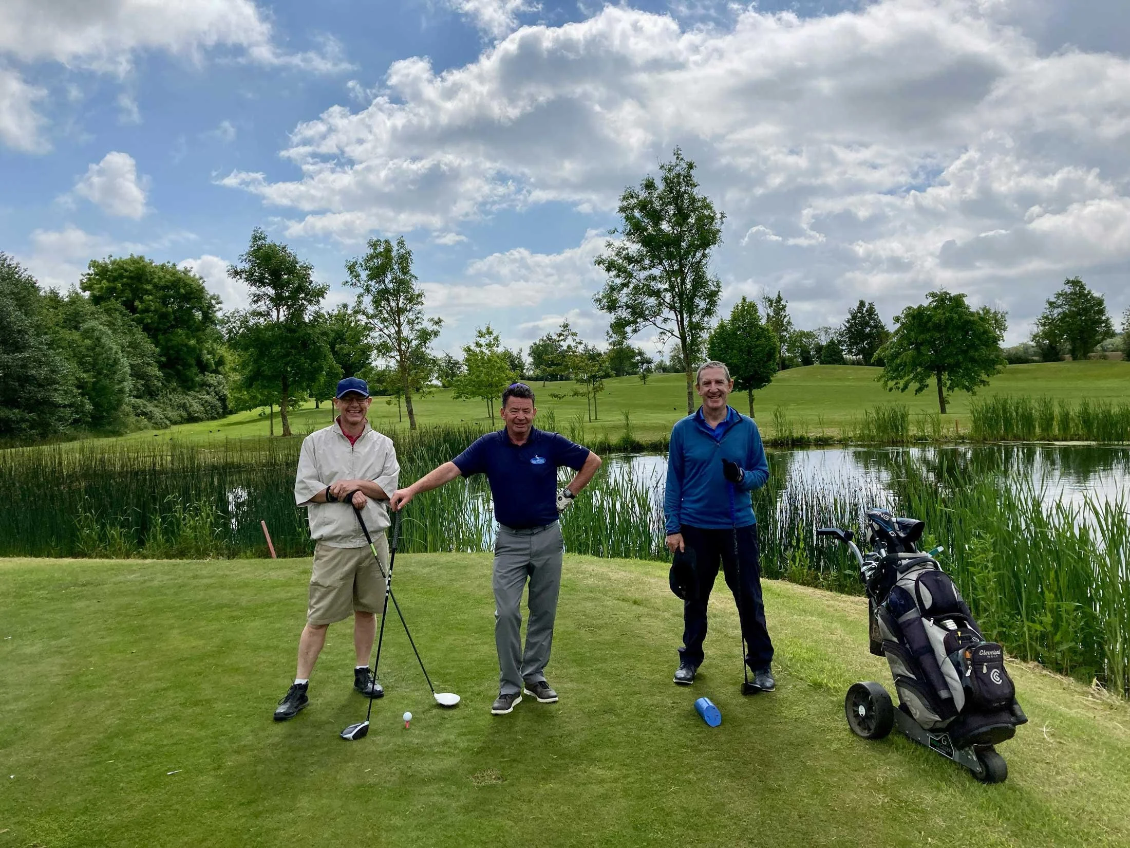 Three men standing on a golf course near a pond, holding golf clubs, with golf bags and a cart nearby, under a partly cloudy sky.