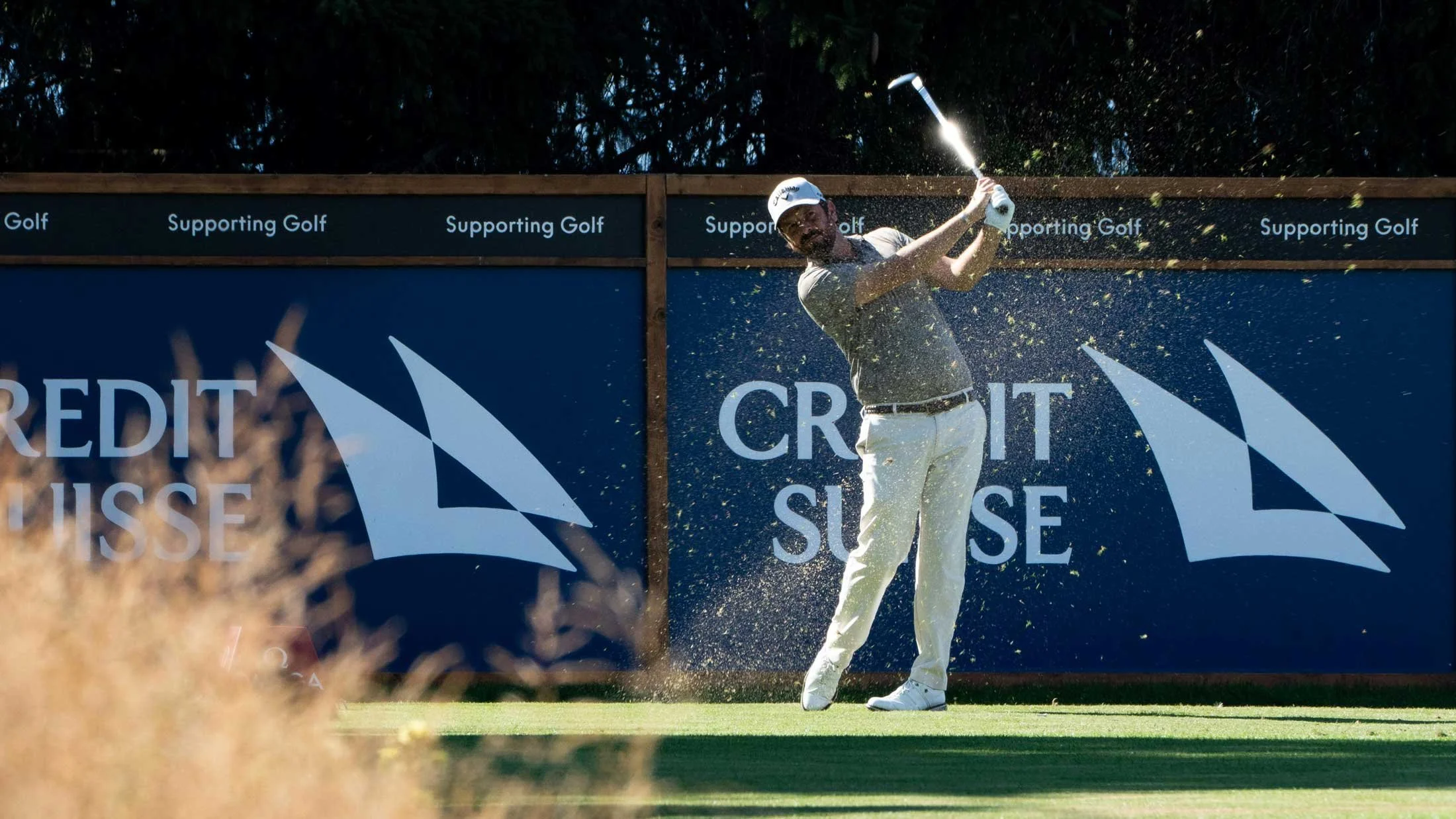 A male golfer swinging a golf club with a golf ball flying through the air. He is on a golf course, wearing white pants, a white cap, and a gray shirt. There is a blue advertising banner behind him with the logos and text about supporting golf and credit Suisse.
