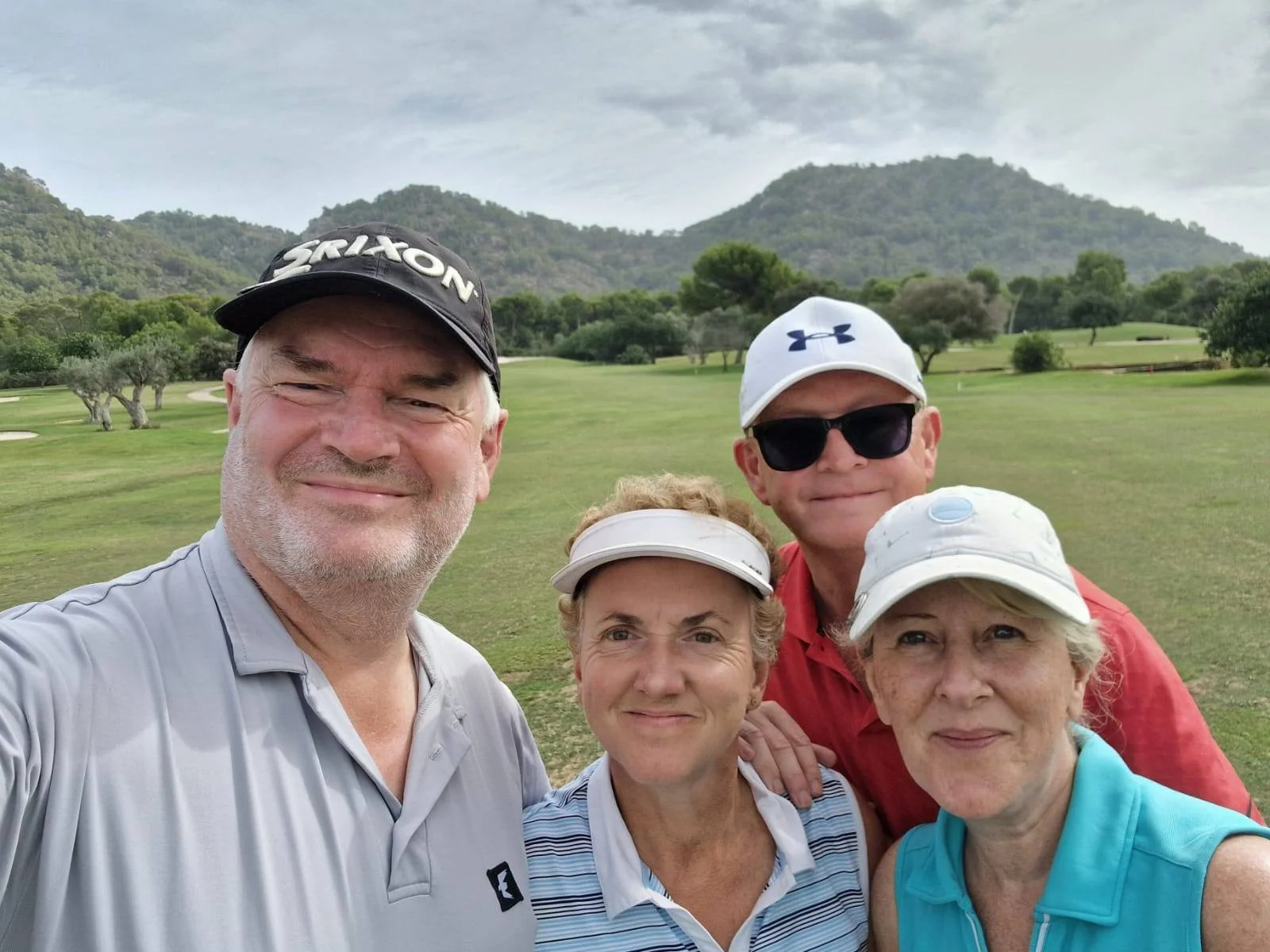 A group of five older adults smiling and posing for a selfie on a golf course with mountains and trees in the background, wearing golf caps and casual sportswear.