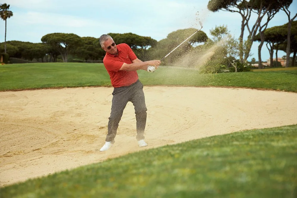 Man in red shirt and gray pants hitting a golf ball from a sand trap on a golf course.