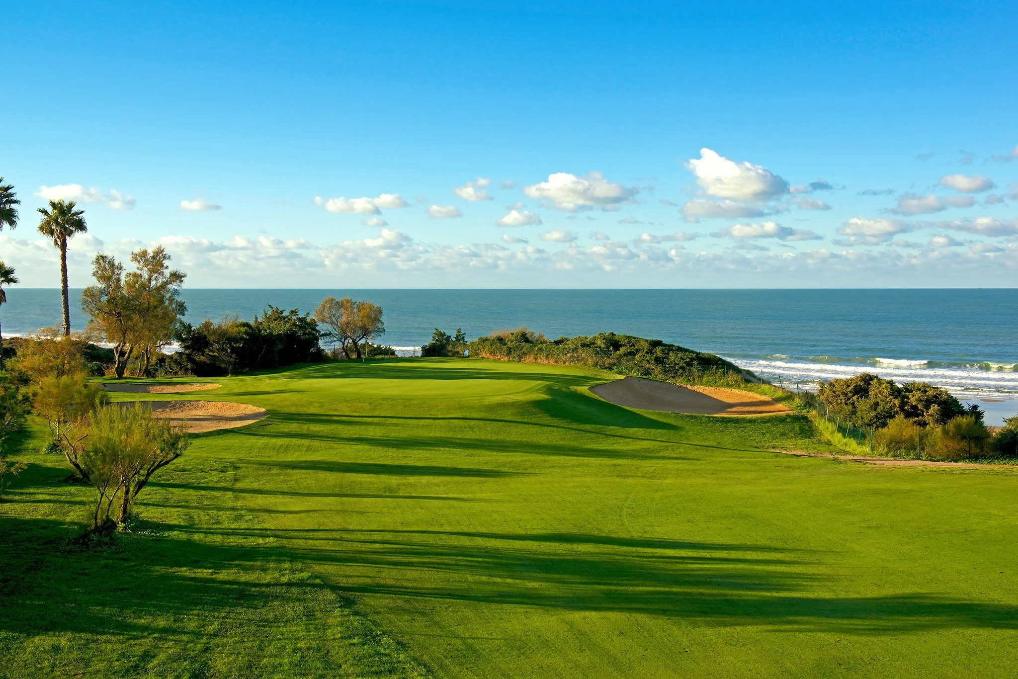 A scenic view of a golf course near the ocean with green grass, trees, and clear blue sky with some clouds.