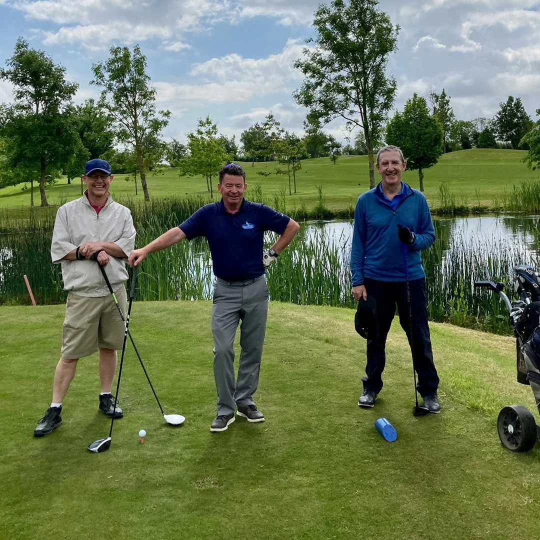Three men standing on a golf course near a pond, holding golf clubs and smiling. They are dressed in casual golf attire with trees and a partly cloudy sky in the background.