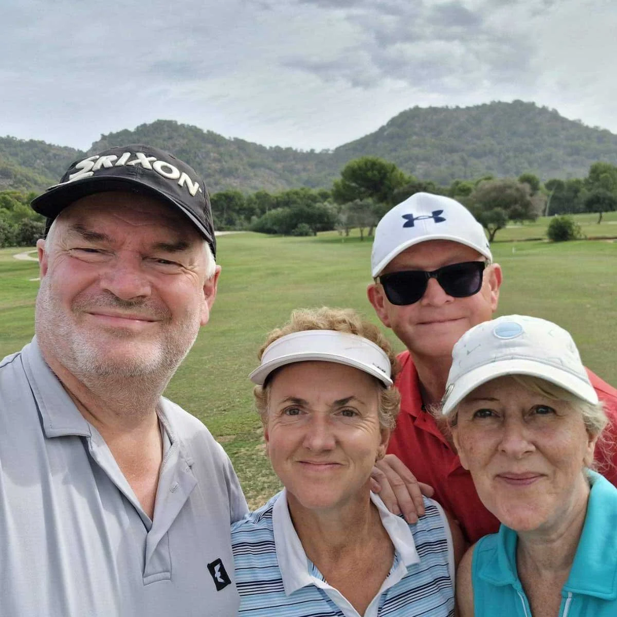 Group of four middle-aged adults posing for a selfie on a golf course with green grass, trees, and hills in the background, wearing golf caps and casual golf attire.