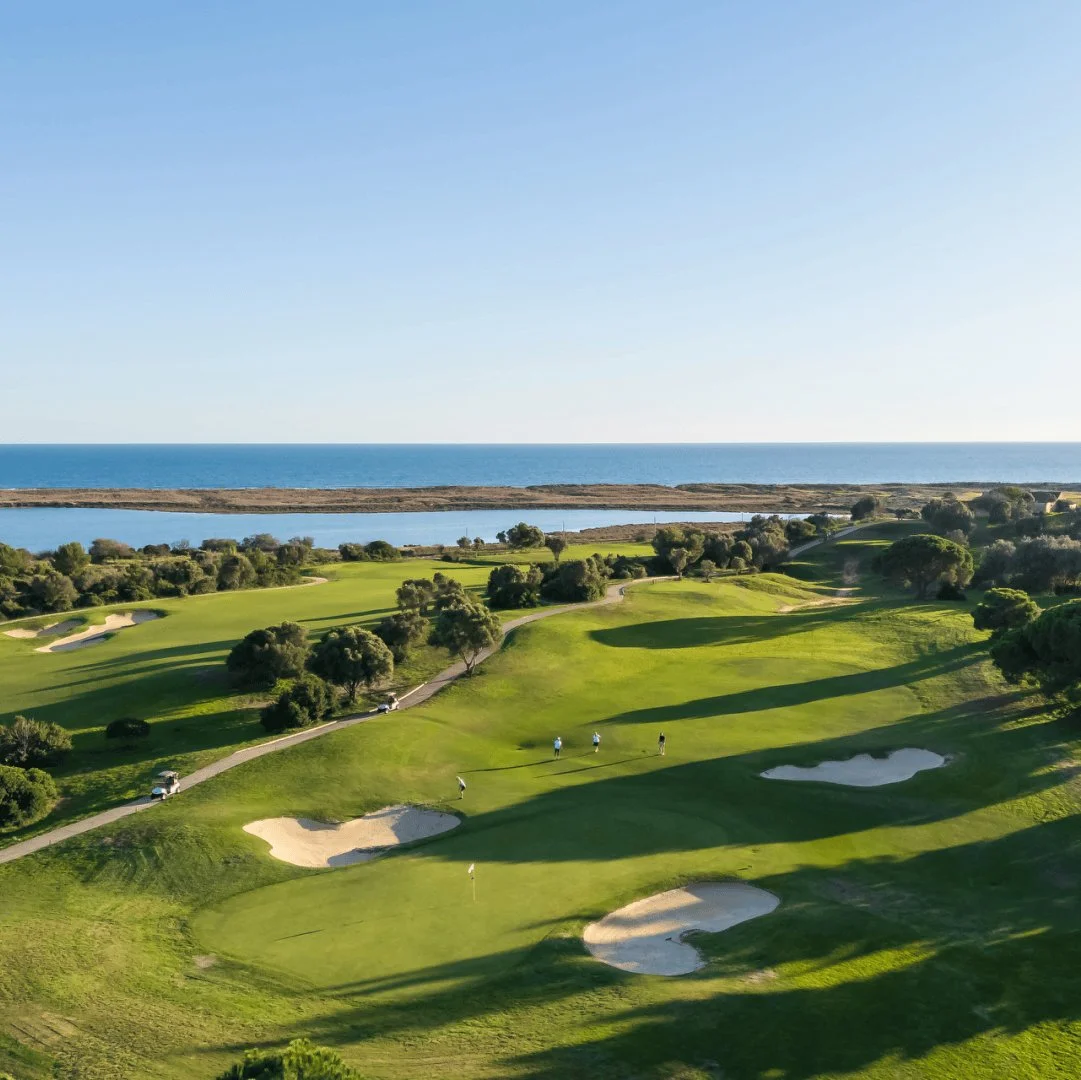 A scenic view of a golf course near the ocean, with green fairways, sand bunkers, trees, and people playing golf during the daytime.