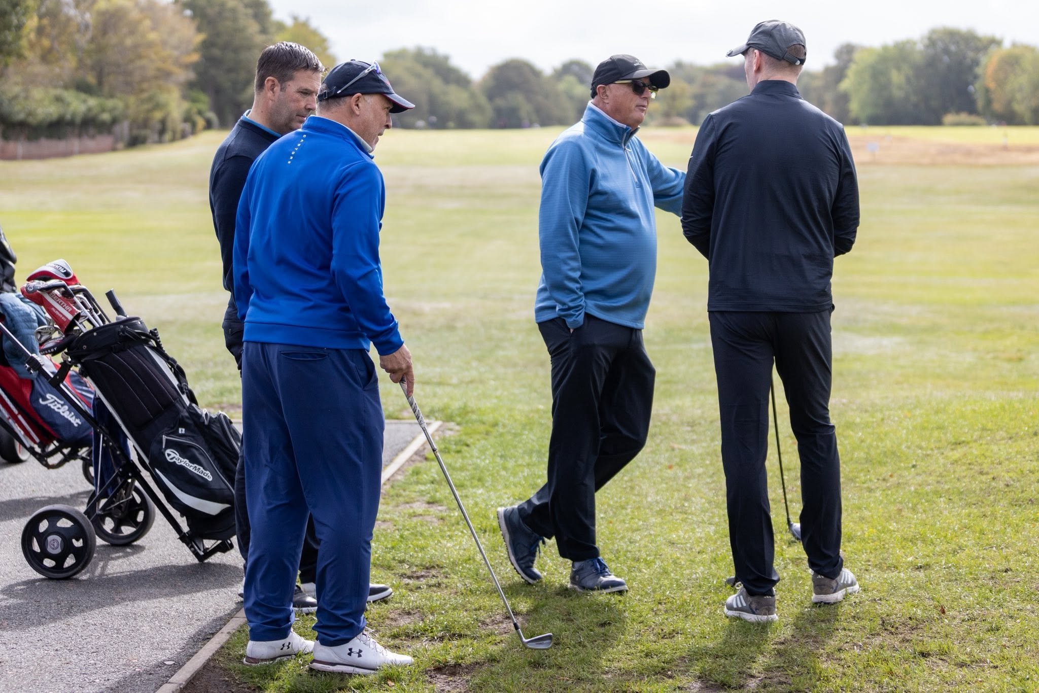 Four men standing on a golf course, two with golf clubs, one in a wheelchair, dressed in golf attire, with golf bags nearby.