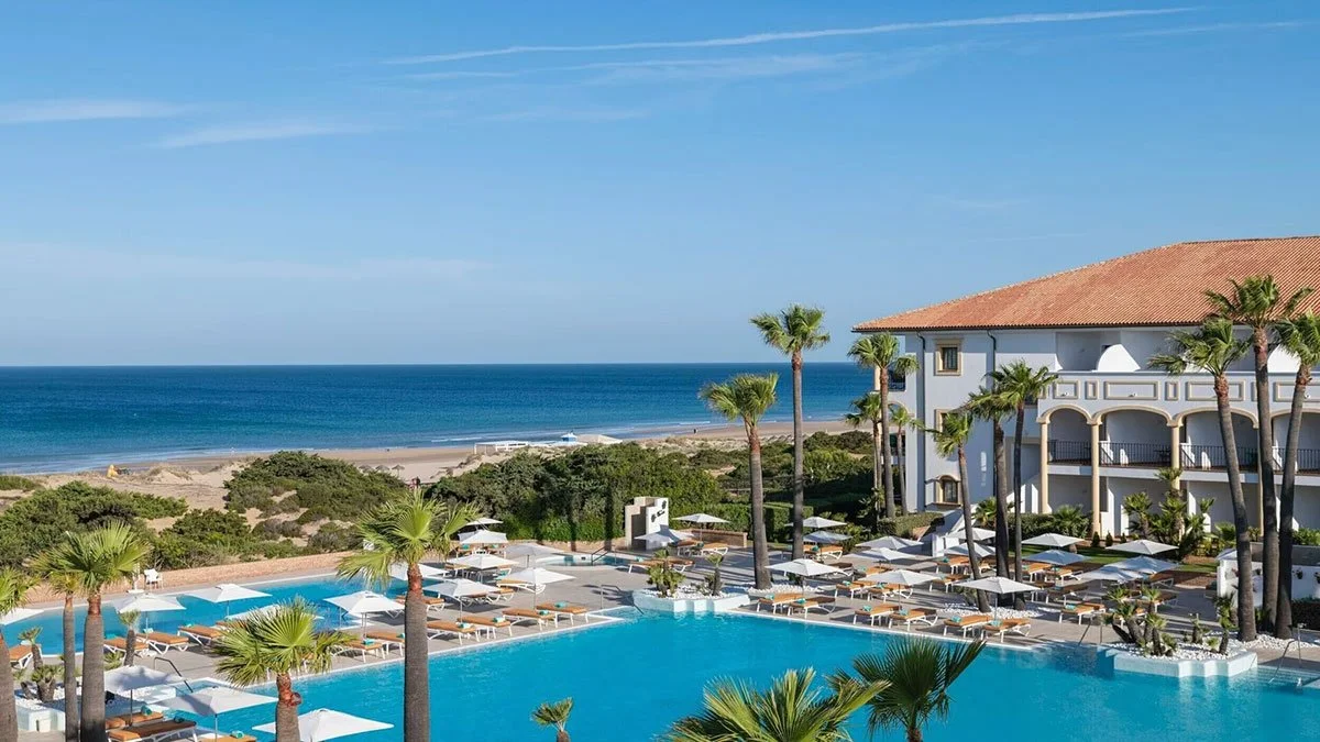 Resort with a large swimming pool, many lounge chairs with umbrellas, palm trees, a building with a terracotta roof, and a view of the ocean in the background under a blue sky.