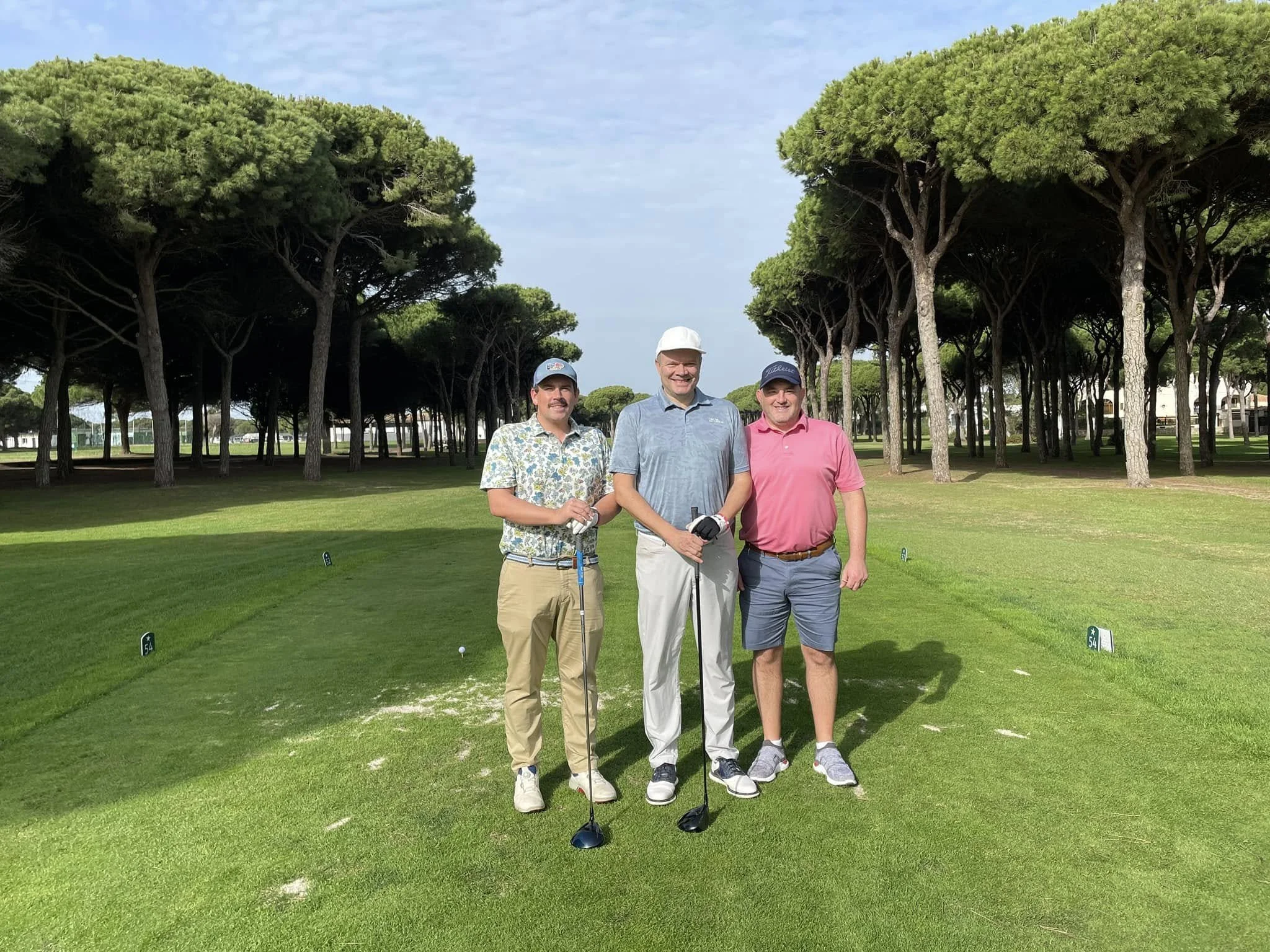 Three men standing on a golf course with trees in the background and golf clubs in their hands.
