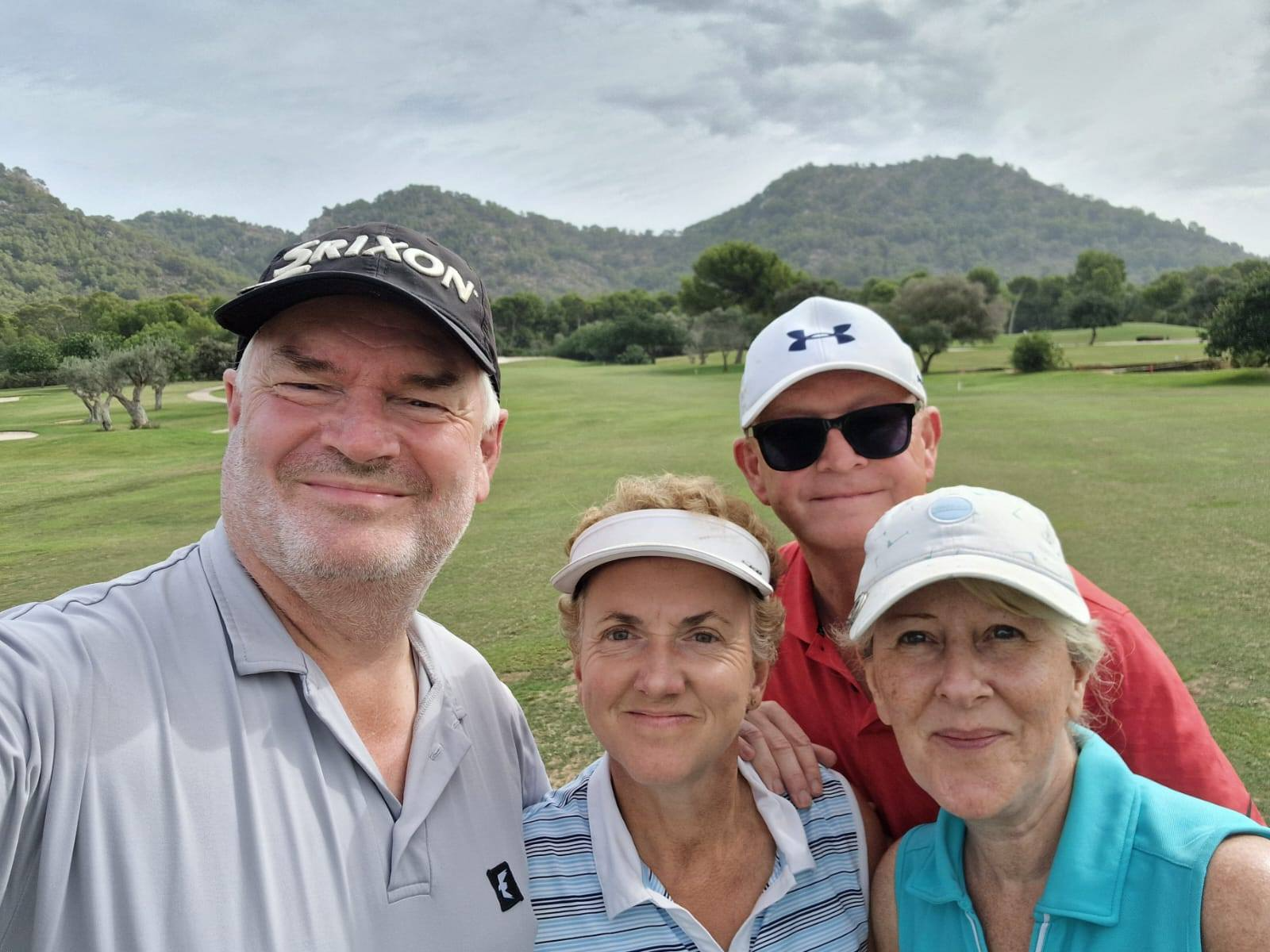 Four people standing on a golf course with hills and trees in the background, smiling at the camera.
