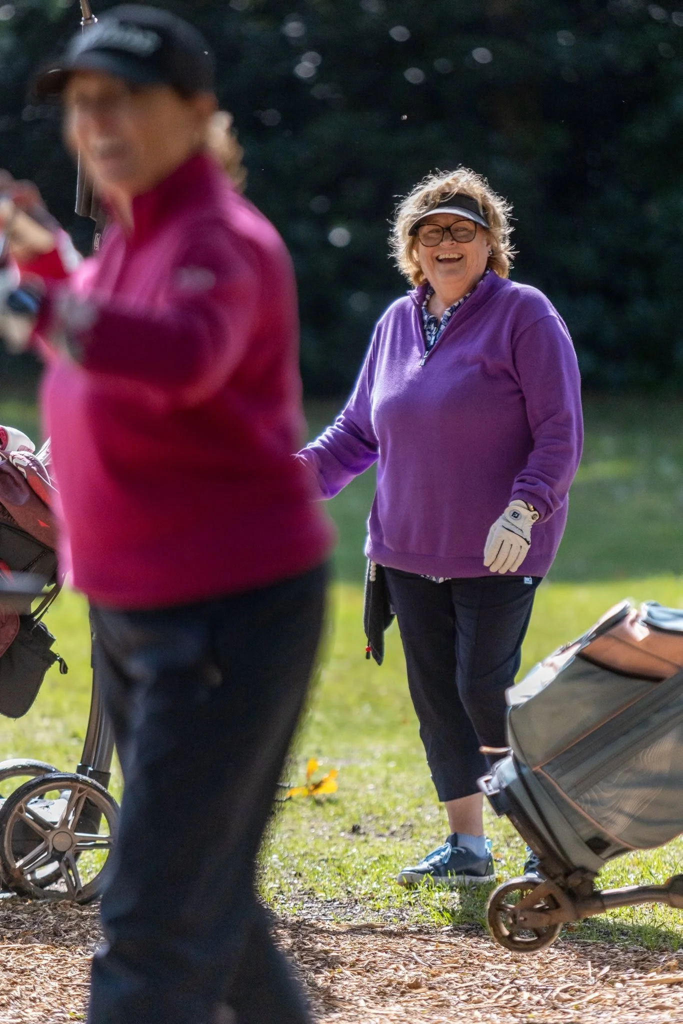A woman with curly hair, glasses, and a purple jacket smiling and standing outdoors next to a stroller. Another person, partially visible, is in the foreground, wearing a pink jacket and a cap, holding a golf club. The scene is set in a grassy park area with sunlight filtering through trees.
