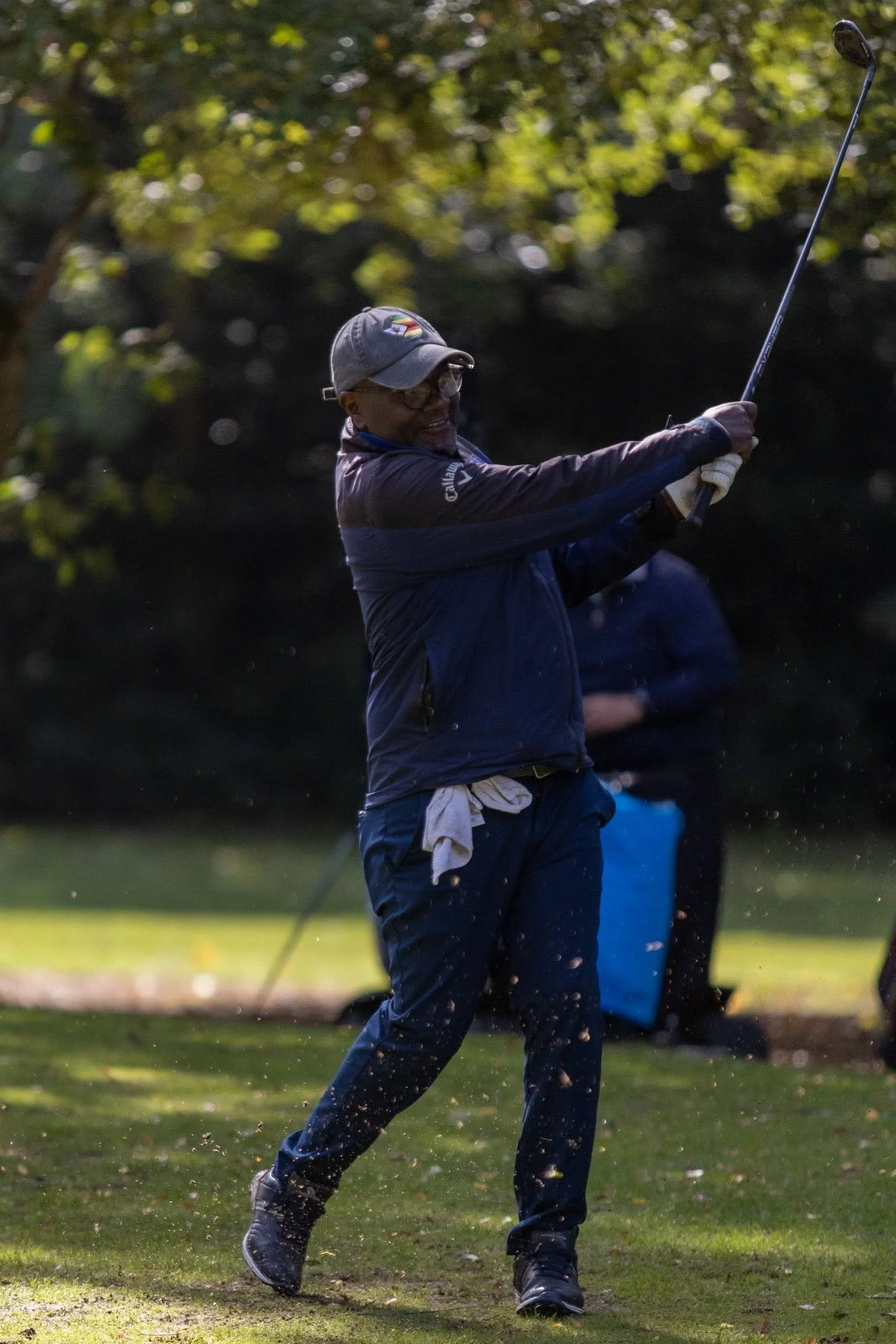 A man in outdoor clothing and a cap swinging a golf club during a game on a golf course, with trees and sunshine in the background.