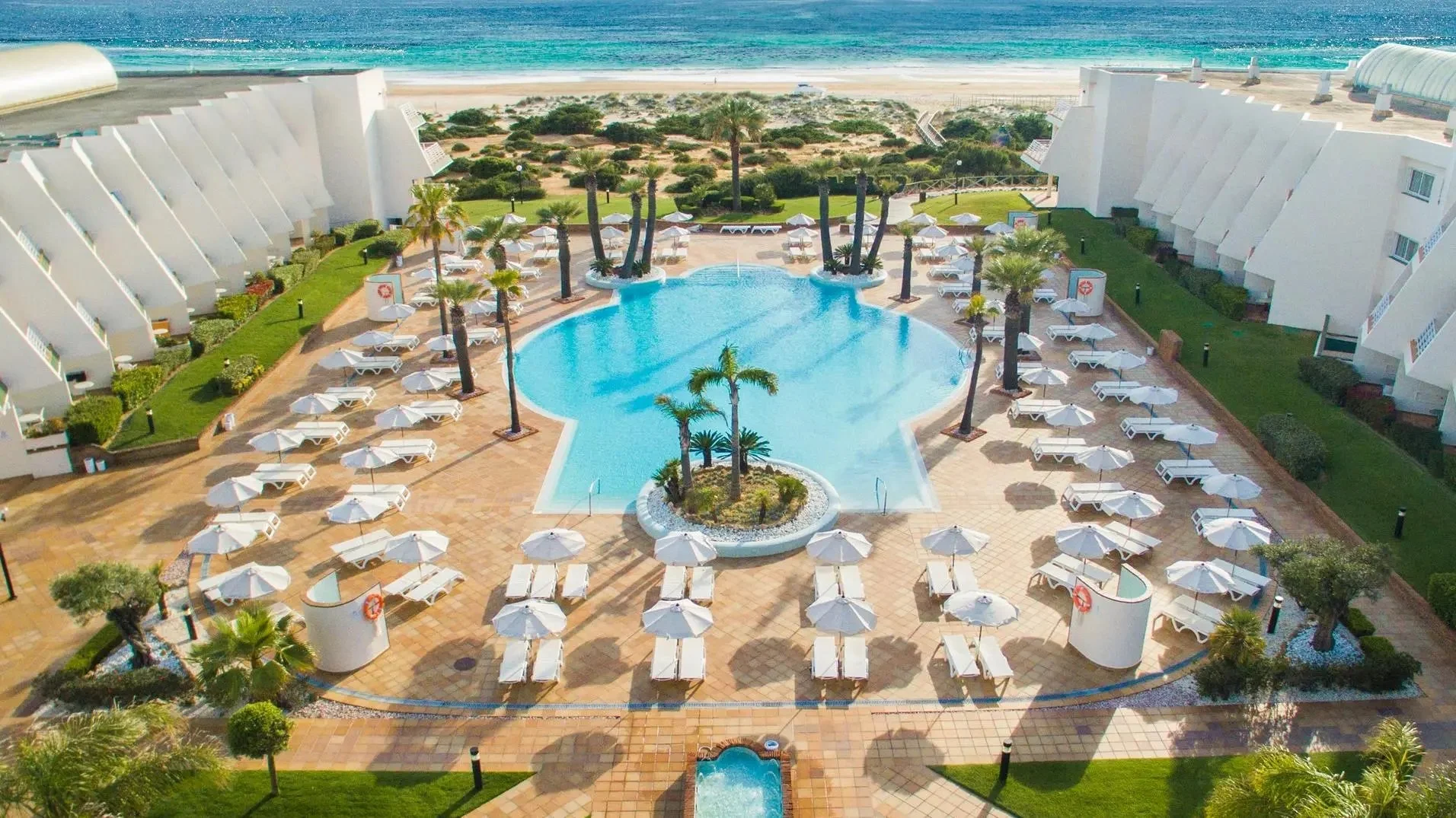 An aerial view of a resort with a large swimming pool surrounded by white lounge chairs and umbrellas, palm trees, well-manicured lawns, and the beach with ocean water in the background.
