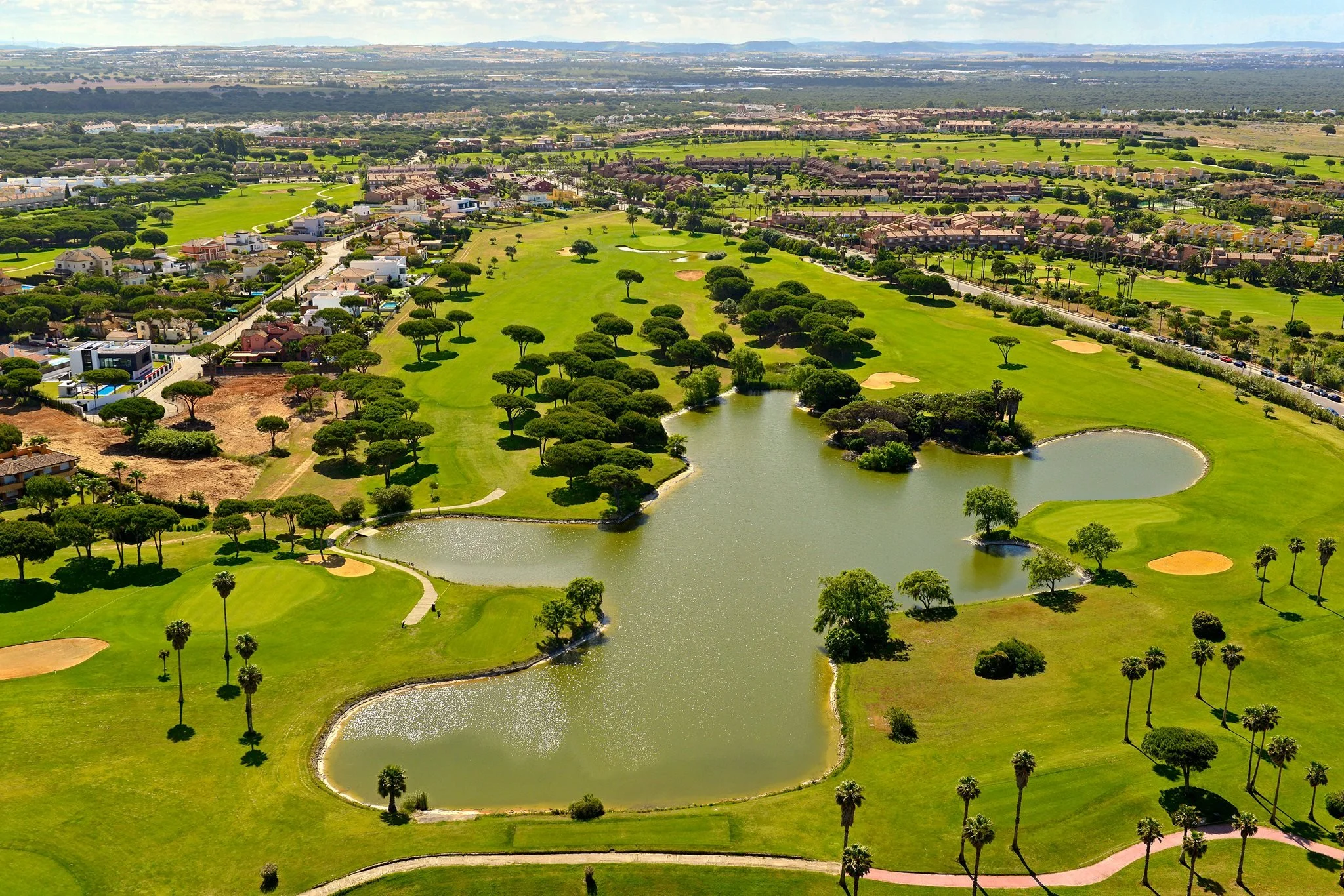 Aerial view of a golf course with green lawns, trees, water hazards, and surrounding residential neighborhoods.