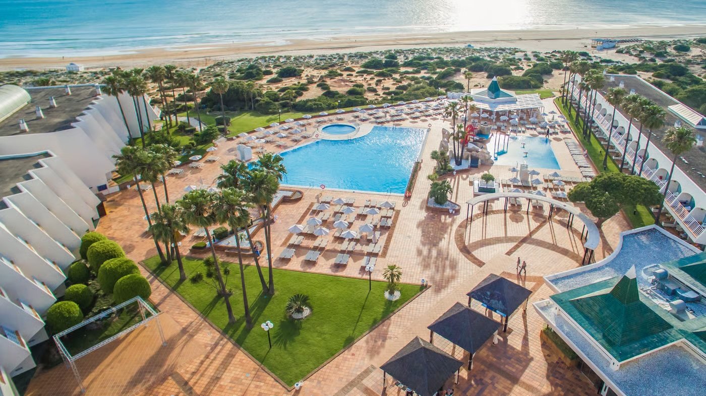 Aerial view of a beachside resort pool area with lounge chairs and umbrellas, palm trees, and a sandy beach with ocean waves in the background.