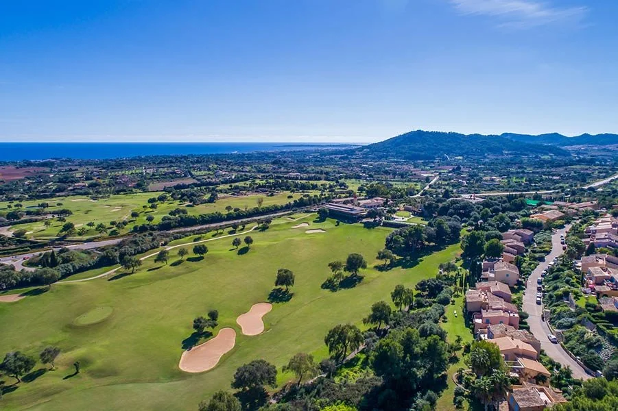 Aerial view of a golf course with sand traps and trees, bordering a residential neighborhood with houses and a winding road, with ocean visible in the distance under a partly cloudy sky.