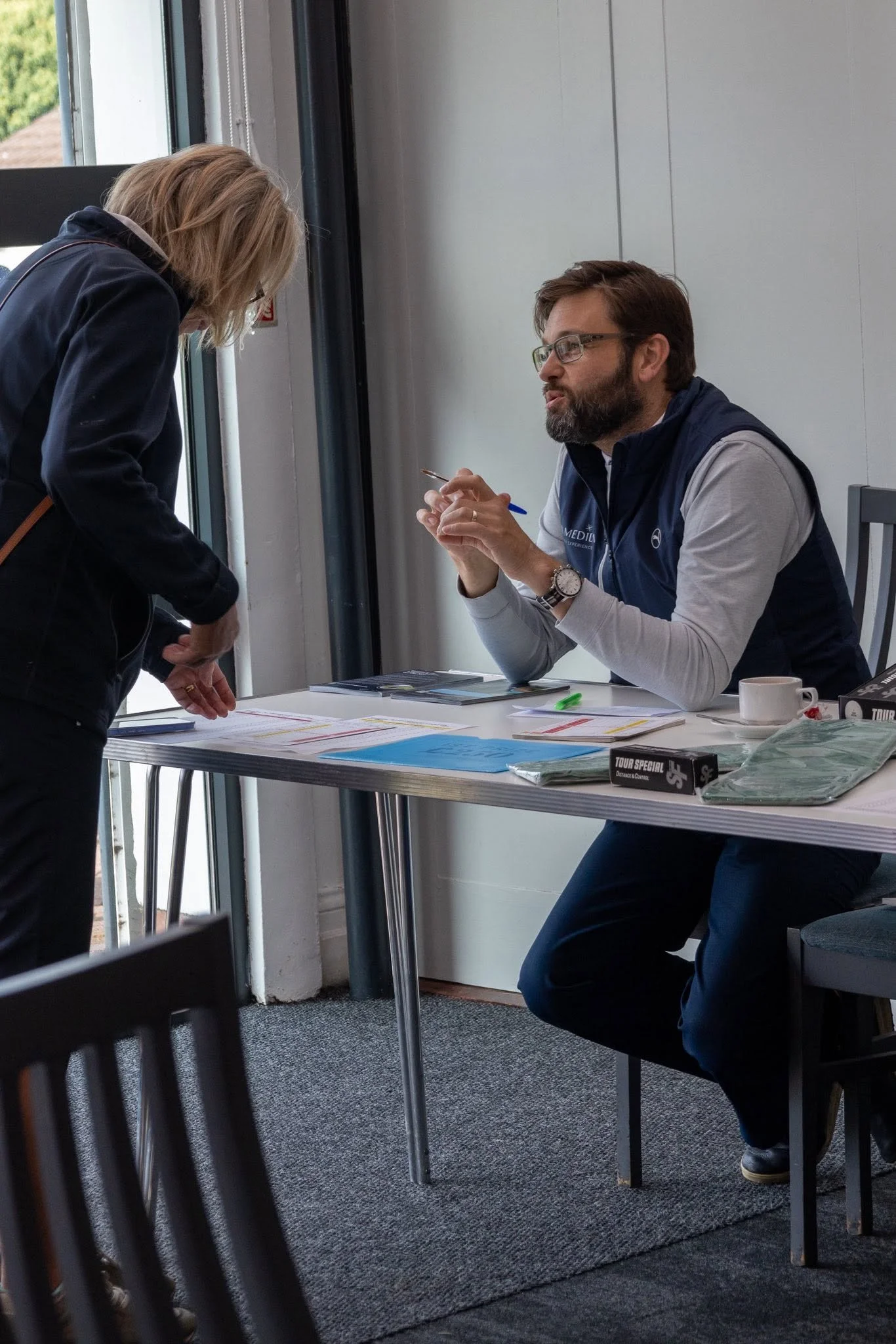 A man with glasses and a beard wearing a vest and gray shirt seated at a table, engaging in conversation with a woman who is leaning over the table, in a room with large windows and gray walls. There are papers, a pen, a mug, and other items on the table.