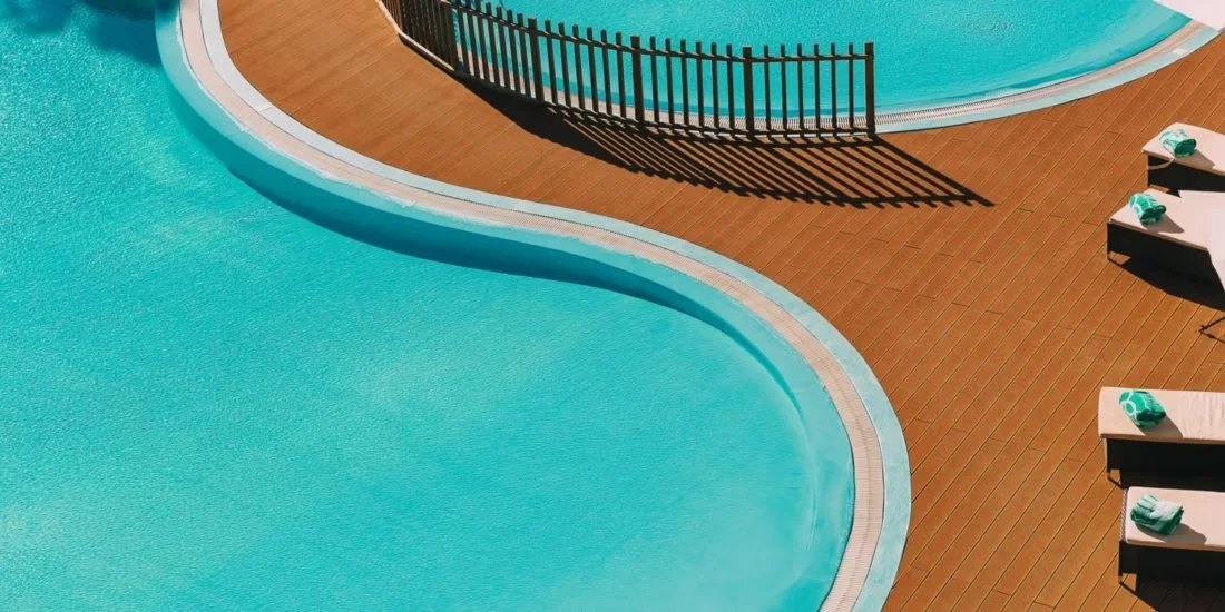 A close-up, top-down view of the curved edge of a bright turquoise swimming pool. A wooden deck with a small fence borders the water, and several lounge chairs with folded teal towels are visible on the right side.