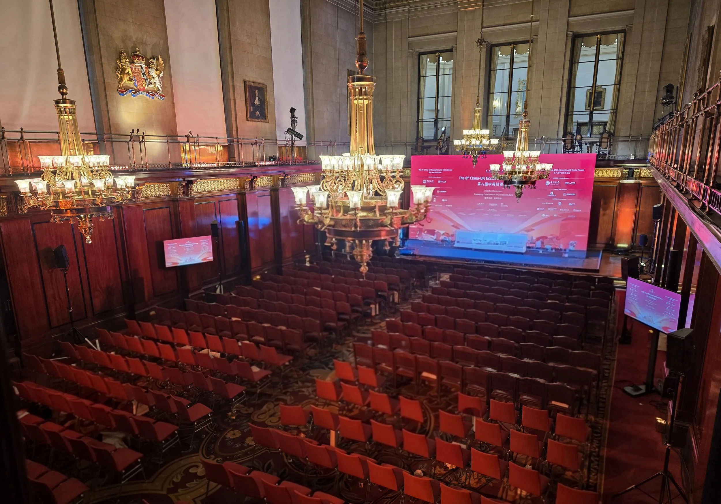 An elegant conference hall with red upholstered chairs arranged theater-style, large chandeliers, wood-paneled walls, and a stage with pink screens displaying event information.