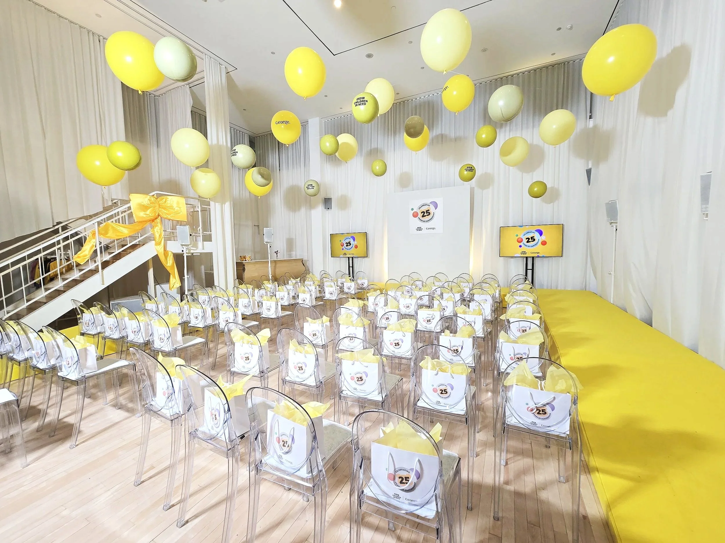A decorated event room with yellow and white balloons, transparent chairs with yellow ribbons, and screens displaying a logo for a 25th anniversary celebration.