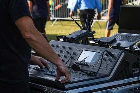 A person adjusting a sound mixing console outdoors with a laptop and audio equipment in the background.