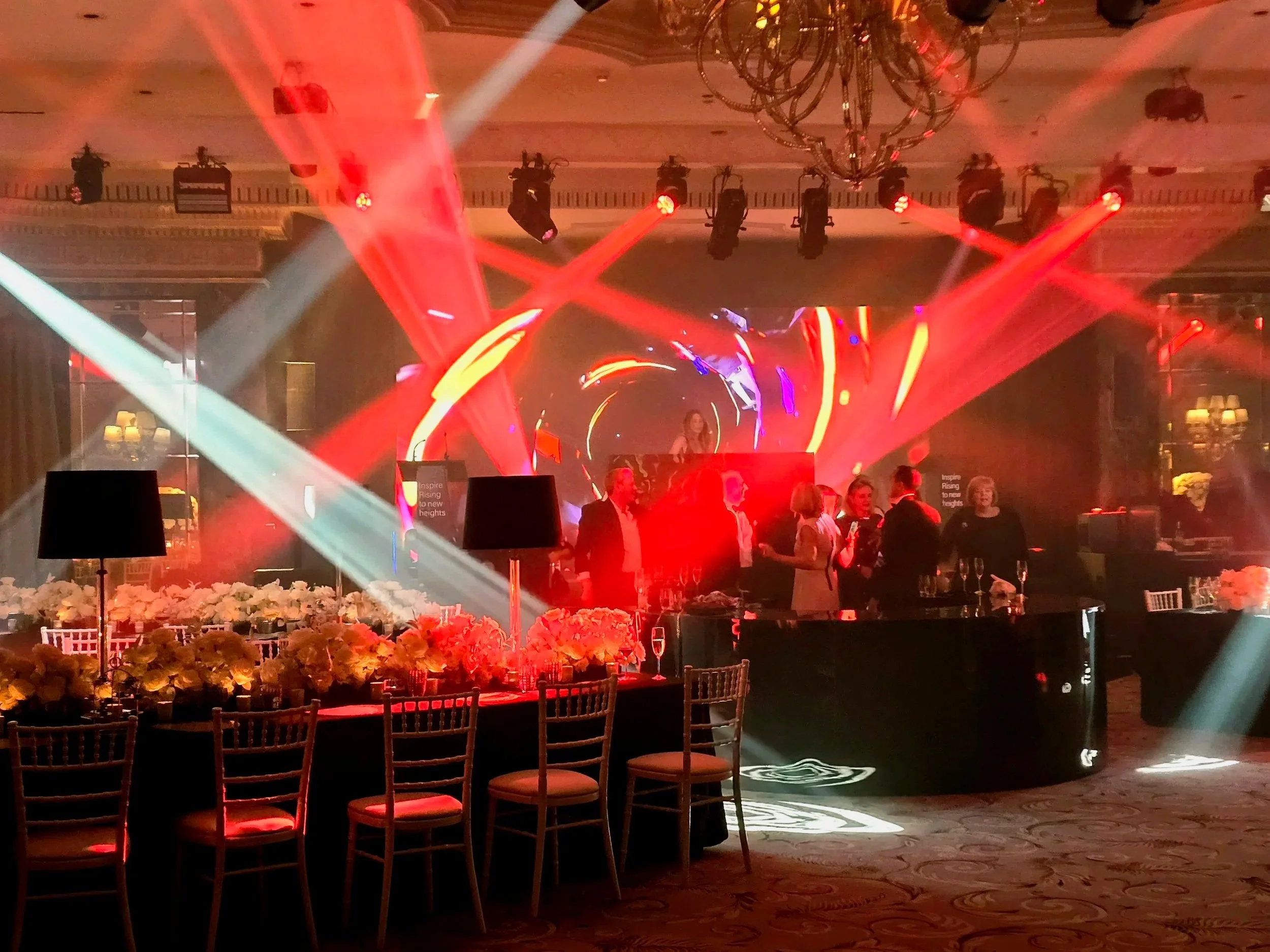 An indoor party venue with colorful stage lighting, a DJ booth with a female DJ, and guests socializing around tables decorated with white flowers and black tablecloths. There are chairs with cushioned seats and a chandelier hanging from the ceiling.