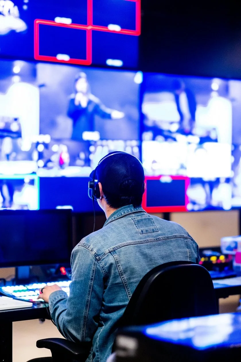 Person in a denim jacket and headphones watching multiple screens in a broadcast or control room.