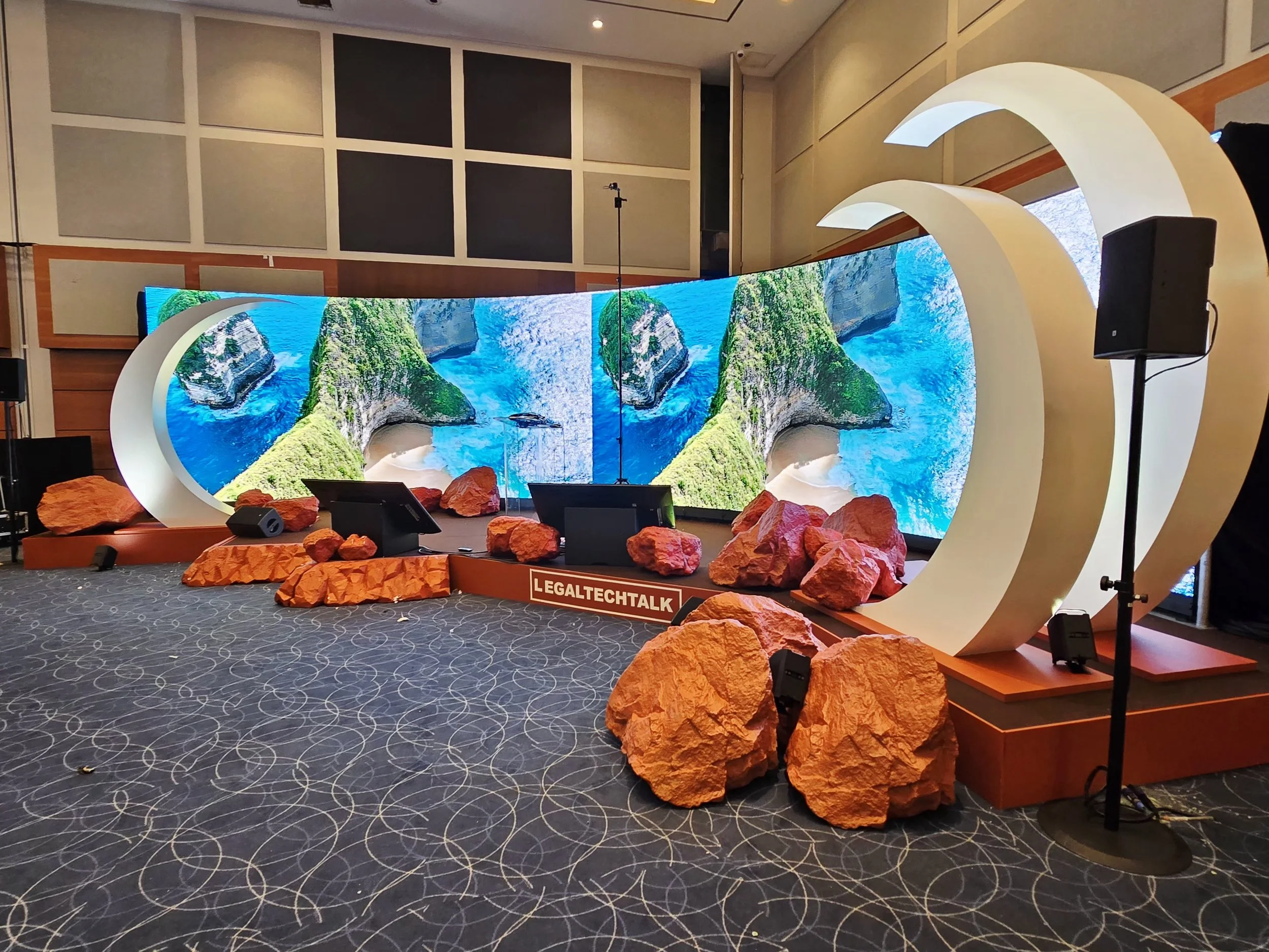 A stage with a large curved screen displaying a tropical landscape with cliffs, ocean, and waterfalls. The stage is decorated with rocks and has a sign that reads "LEGALTECHTALK." There are speakers set up on stands.