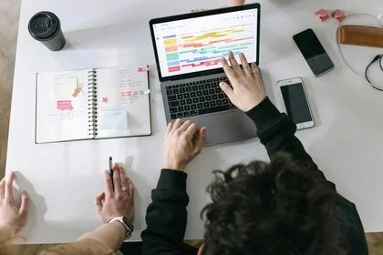 Person working on a color-coded digital planner on a laptop, with a notebook, coffee cup, phone, wallet, glasses, and another smartphone on a white desk.
