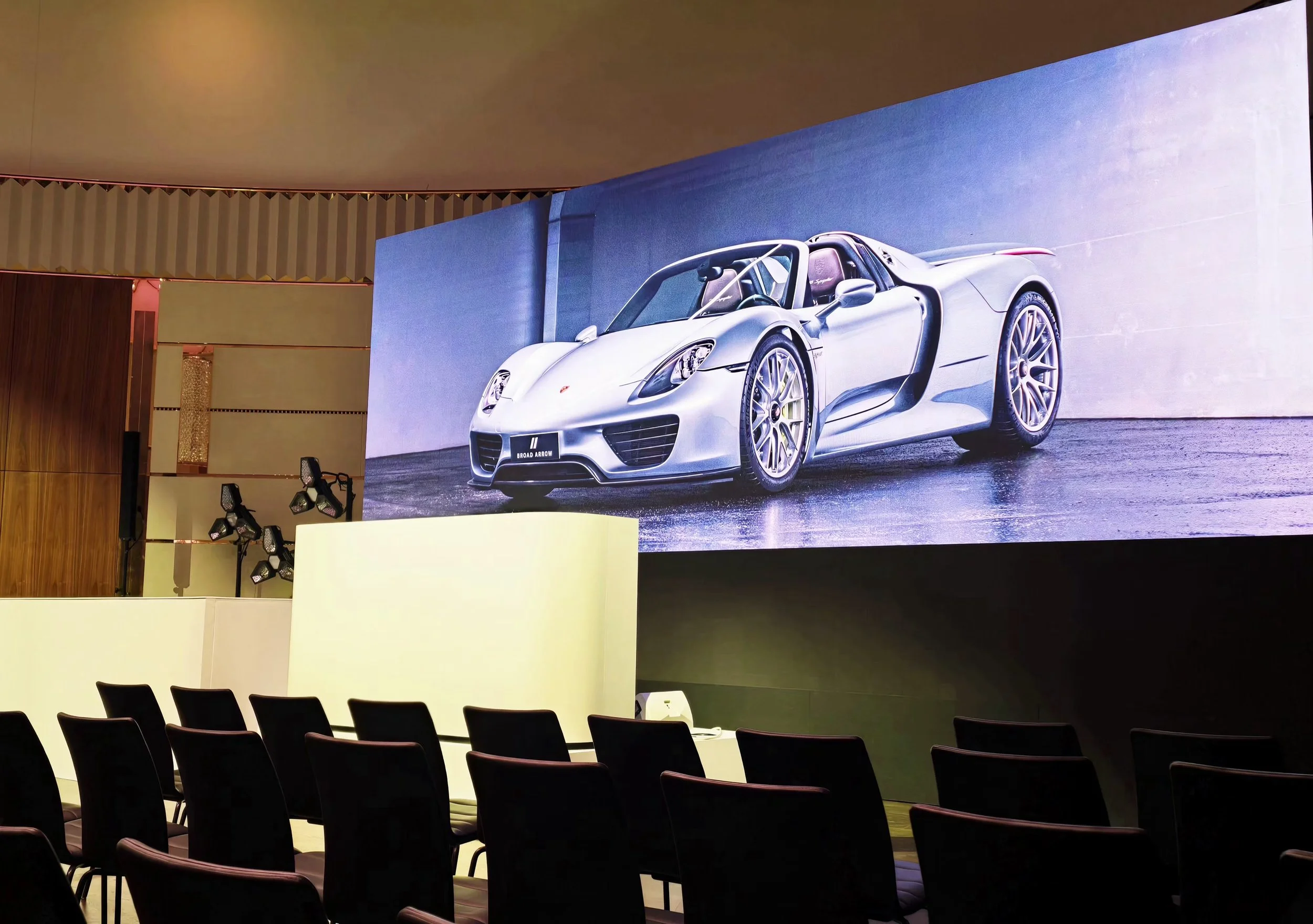 A large presentation screen displays a silver sports car in an auditorium setting with chairs in the foreground.