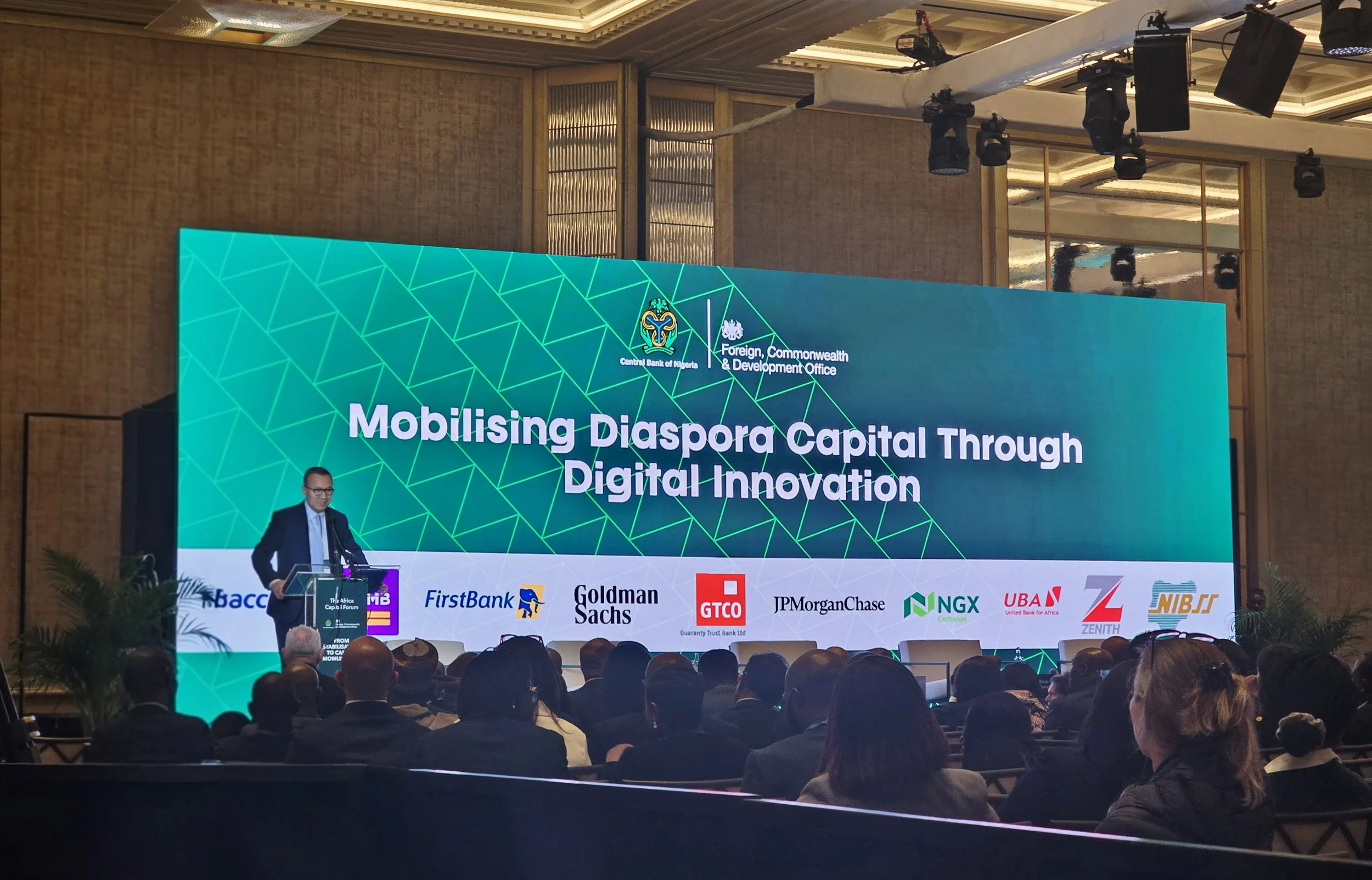 A man in a dark suit and glasses stands at a podium giving a speech in front of a large screen displaying the title 'Mobilising Diaspora Capital Through Digital Innovation' at a conference. The audience is seated facing the stage, and the room has a decorated ceiling and wall panels.