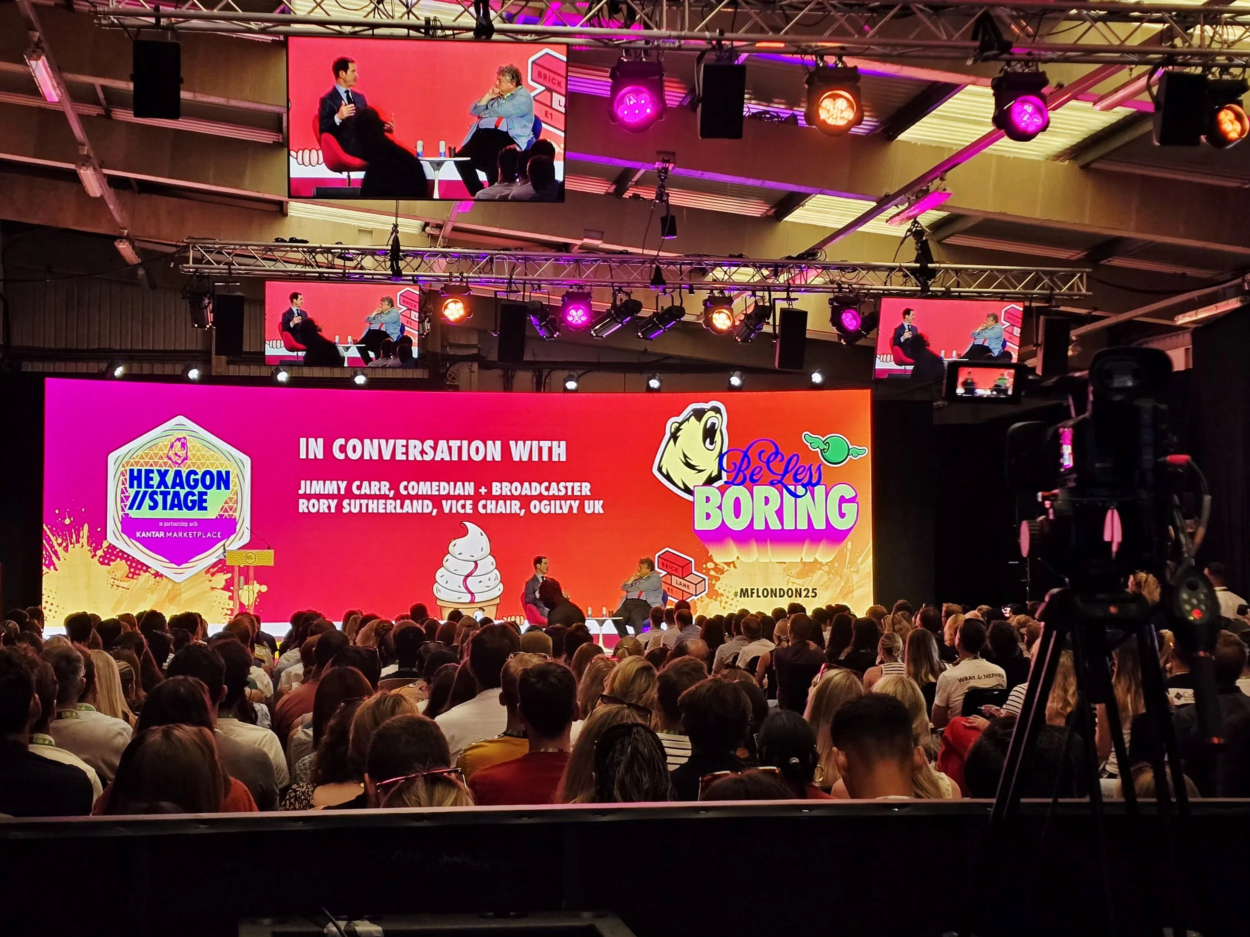A large conference stage with a bright pink and orange backdrop displays the title 'In Conversation With Jimmy Carr, Rory Sutherland, Vice Chair, Ogilvy UK' and event branding for Hexagon/Stage and Be Less Boring, with an audience seated facing the stage. There are several large screens showing the ongoing discussion, with bright stage lighting and a camera on a tripod capturing the event.