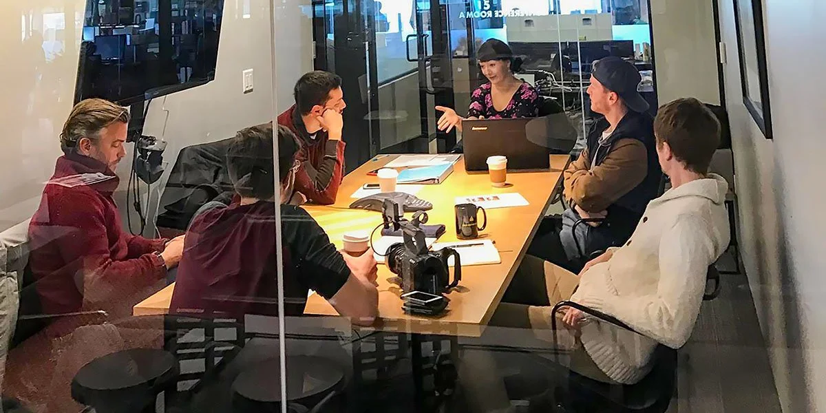 A group of seven people sitting around a rectangular conference table in a modern office, engaged in discussion, with cameras, notebooks, and coffee cups on the table.
