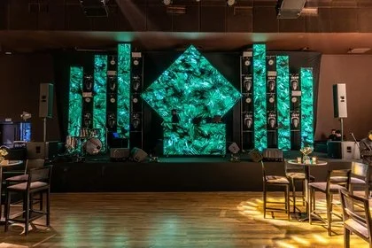 Event stage with large LED screens displaying green abstract patterns, surrounded by speakers and DJ equipment, with tables and chairs in the foreground.