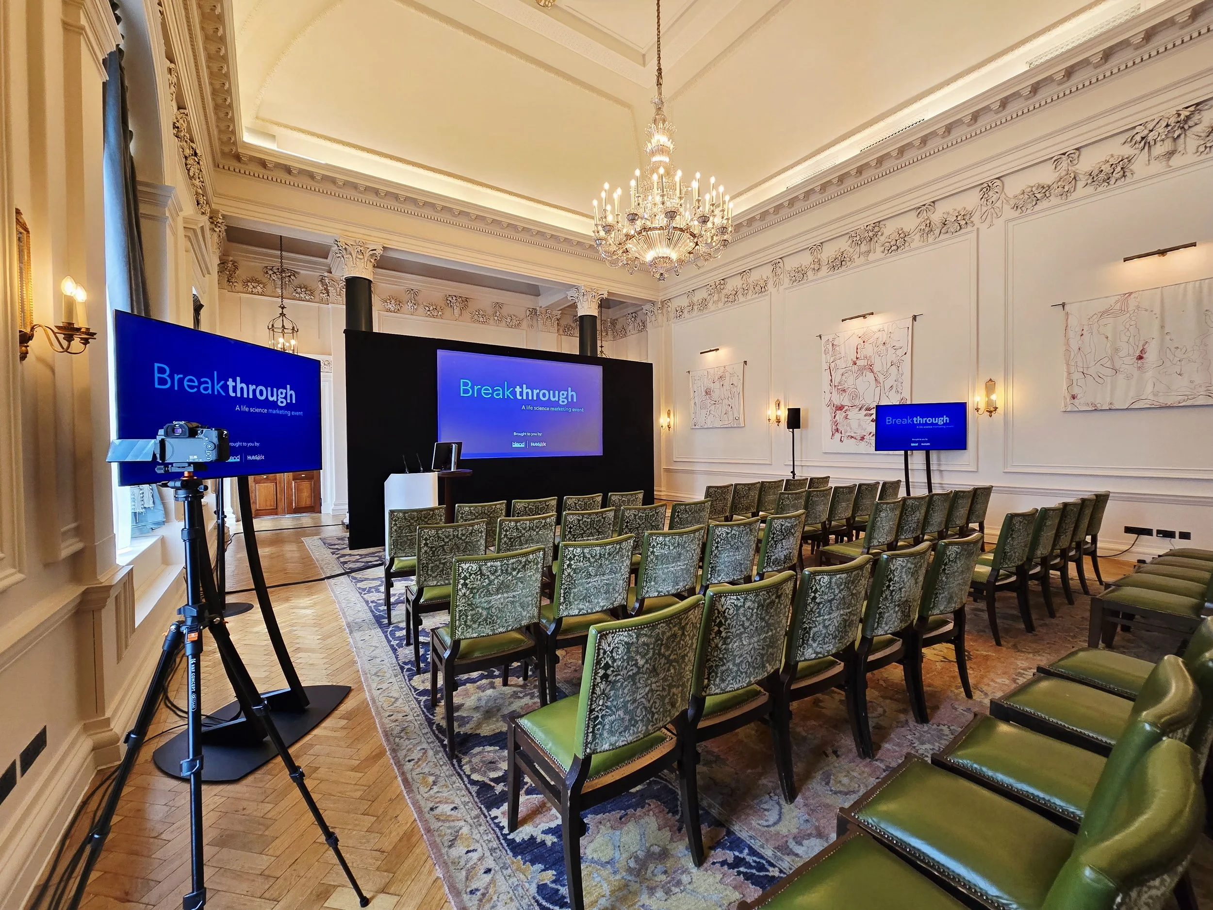 An elegantly decorated conference room with vintage green patterned chairs arranged in rows facing a black stage with two large screens displaying 'Breakthrough'. The room has ornate white walls with intricate moldings, a large crystal chandelier hanging from the ceiling, and artistic wall hangings. A camera on a tripod is set up at the side.