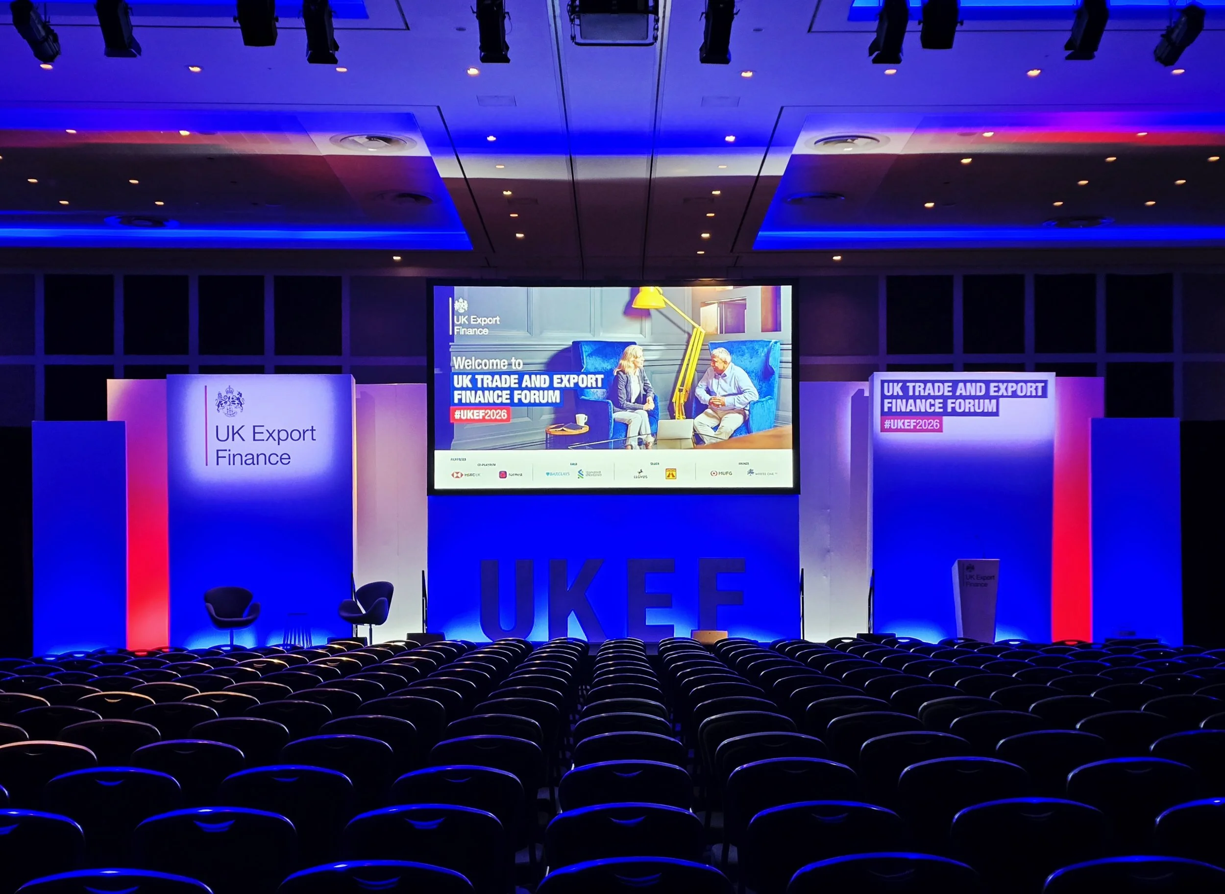 Empty conference hall with rows of chairs facing a stage at the UK Trade and Export Finance Forum, illuminated with blue and purple lighting, large screens displaying event information, and two chairs on stage.
