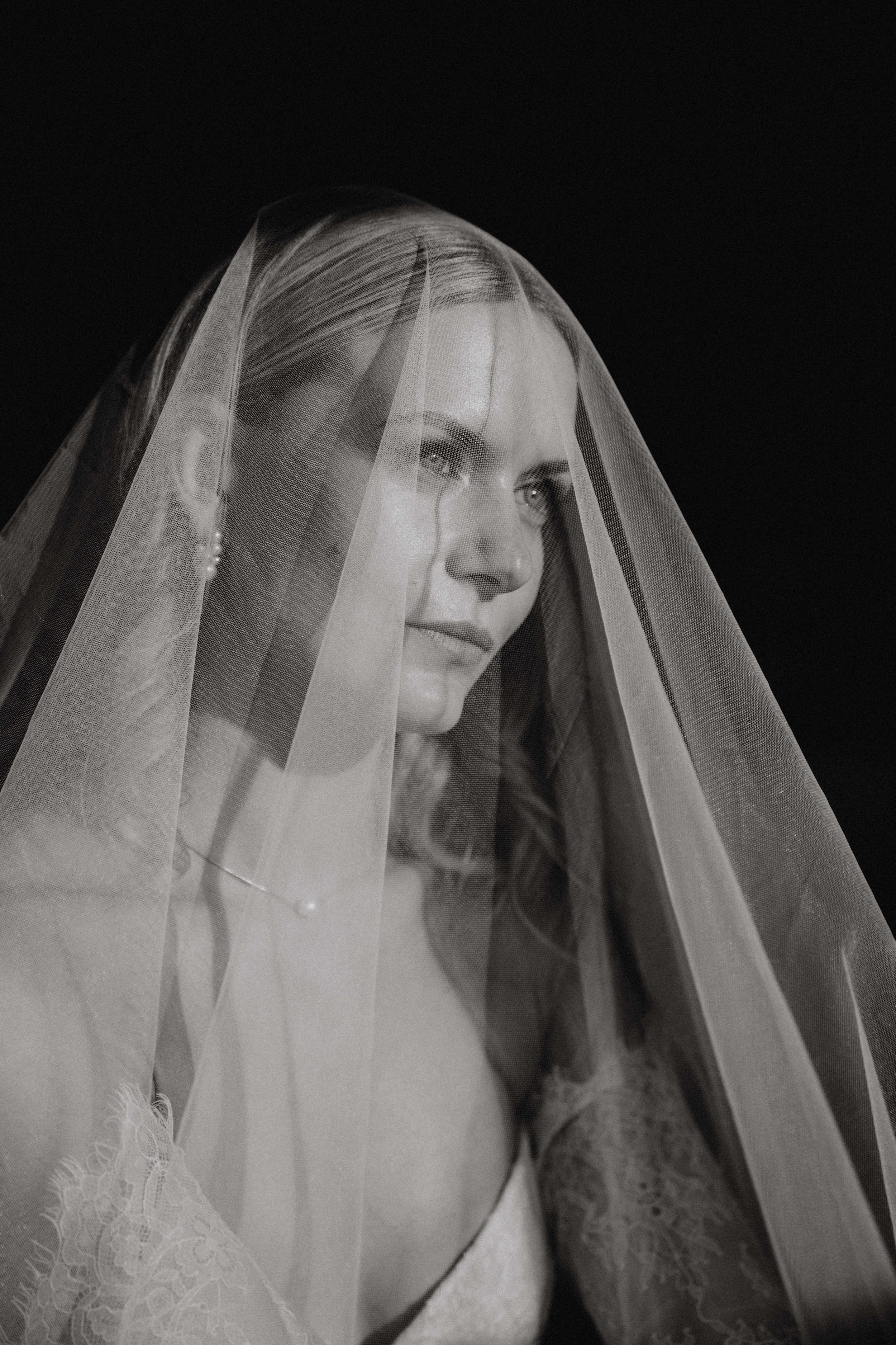 A black and white photograph of a woman with a veil covering her head, looking to her left with a solemn expression.