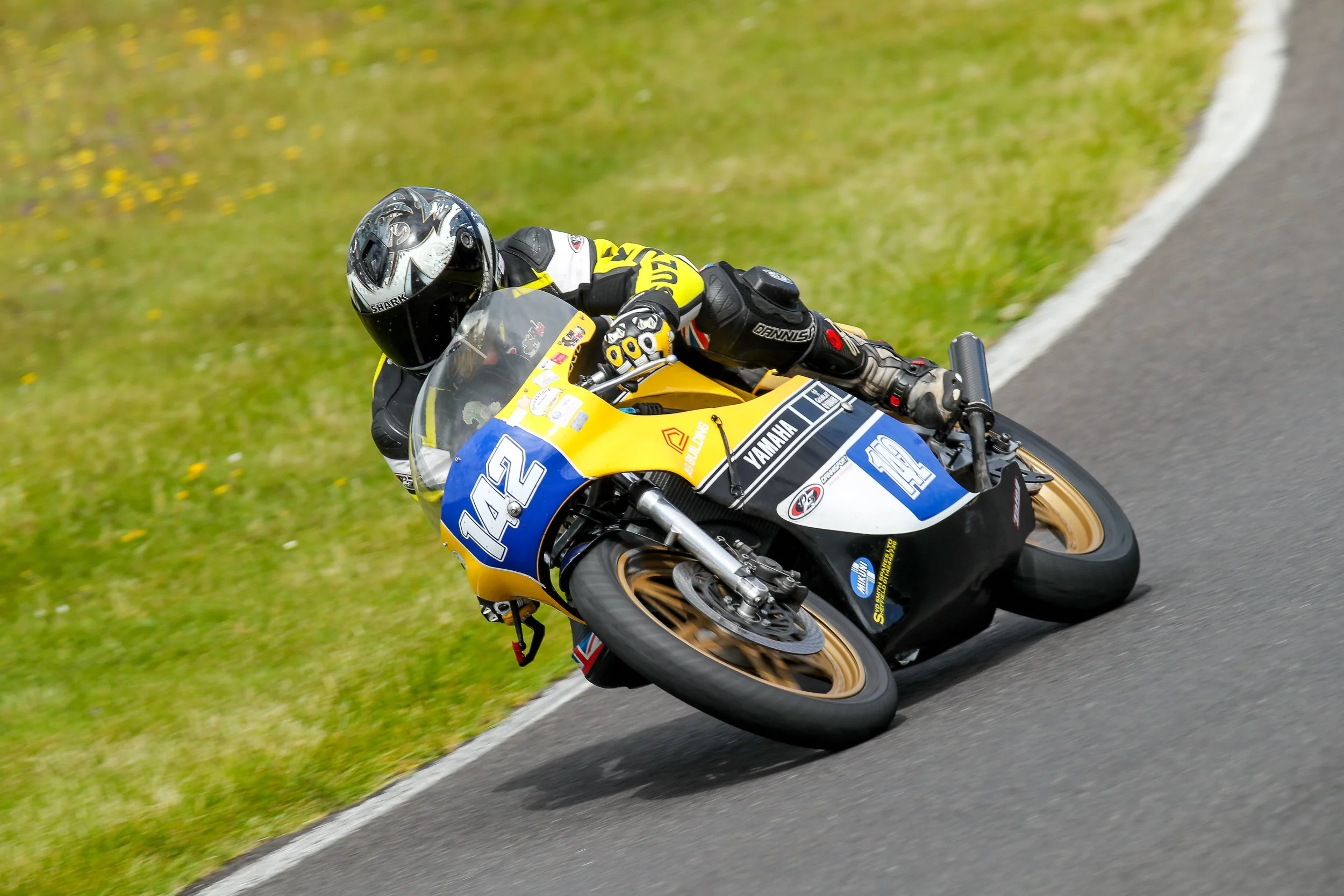 Motorcycle racer leaning into a turn on a race track, wearing black helmet and racing gear, motorcycle number 142 with yellow, blue, and black livery, Yamaha branding, and various sponsorship decals.