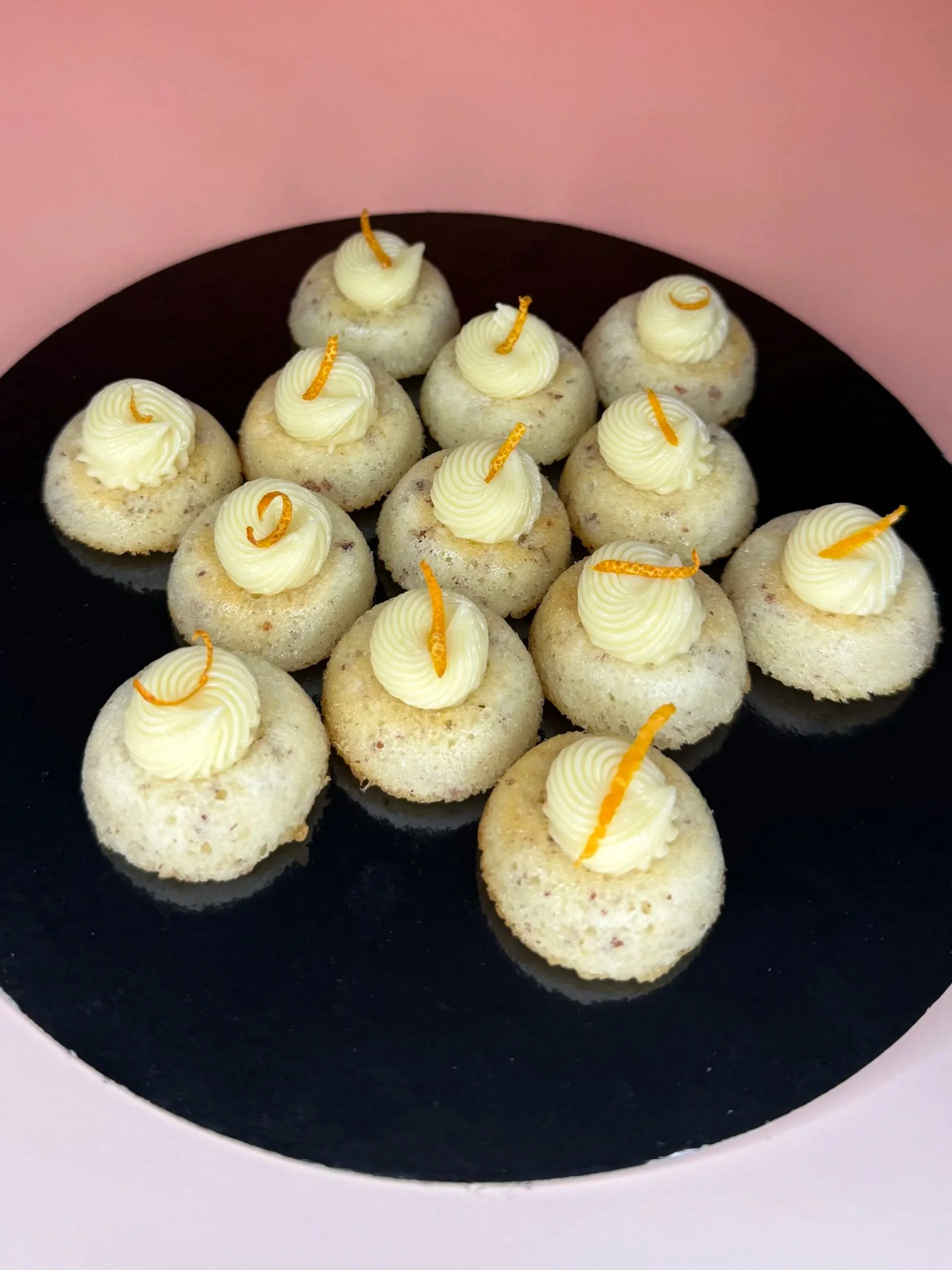 Petits gâteaux ronds garnis de crème blanche et décorés de zestes d'orange confits sur un plateau noir, avec un fond rose.