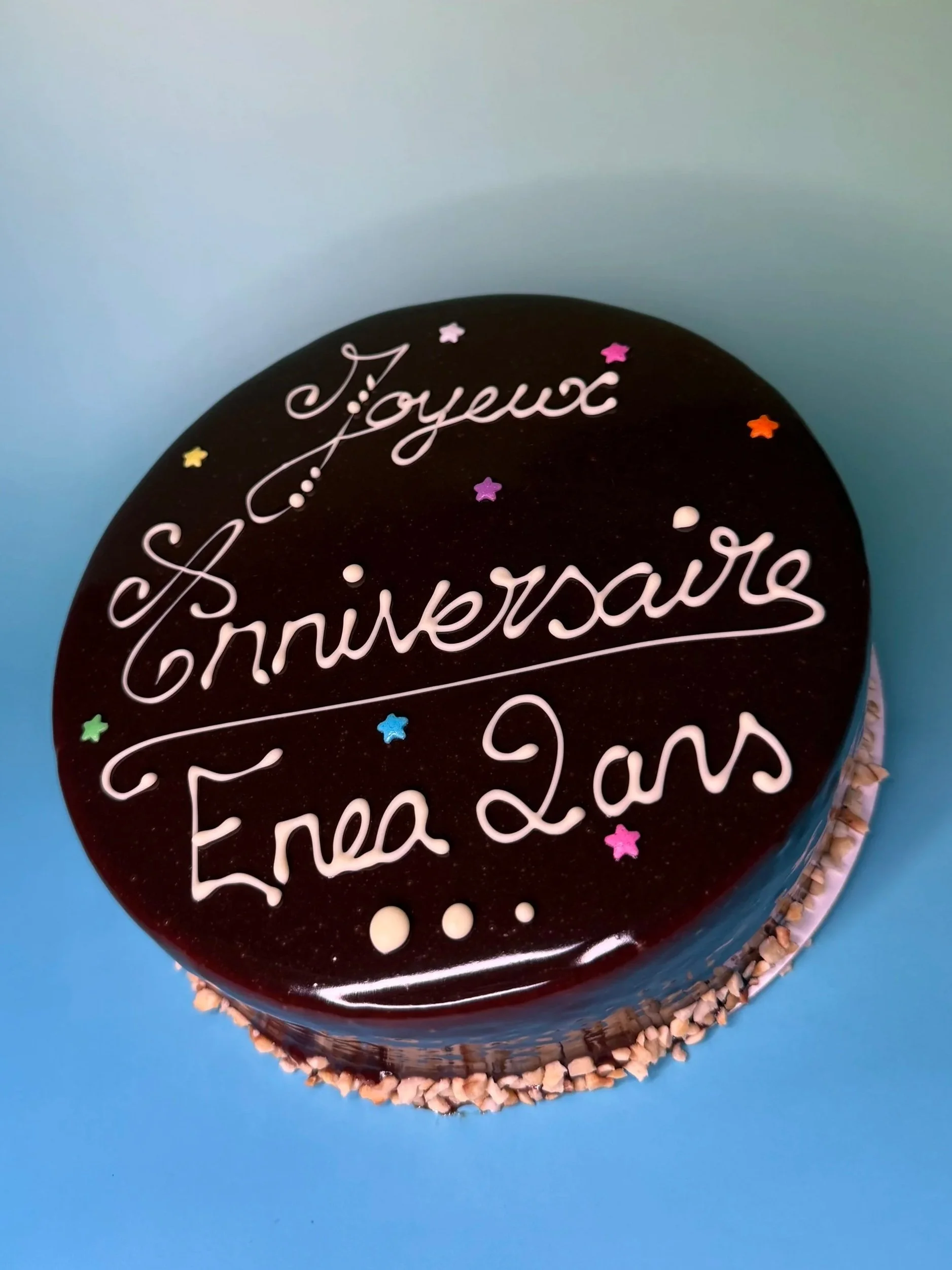 Gâteau rond au chocolat avec des décorations de petites étoiles colorées. Écriture en pâte à sucre disant 'Joyeux Anniversaire Eva 2 ans'.