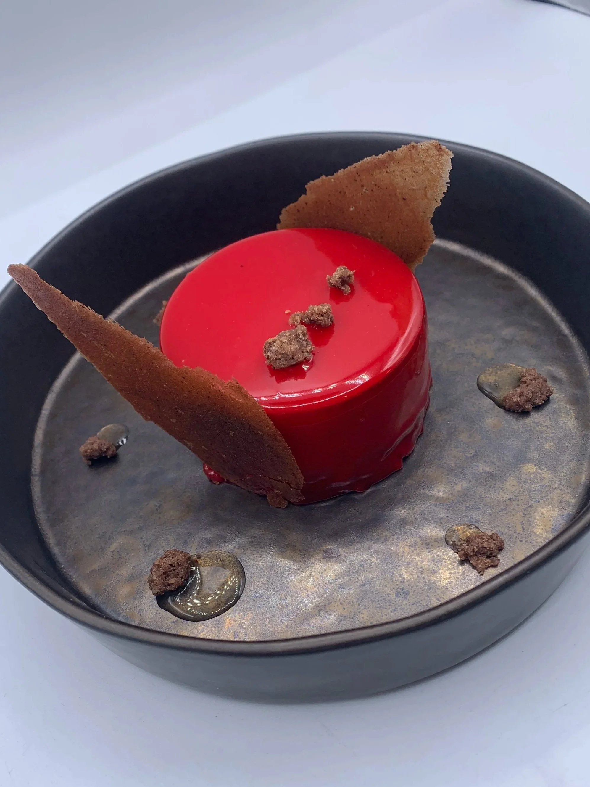 Gâteau miroir rouge avec décoration croustillante, servi dans un plat noir, avec des morceaux de crumble et de la sauce.