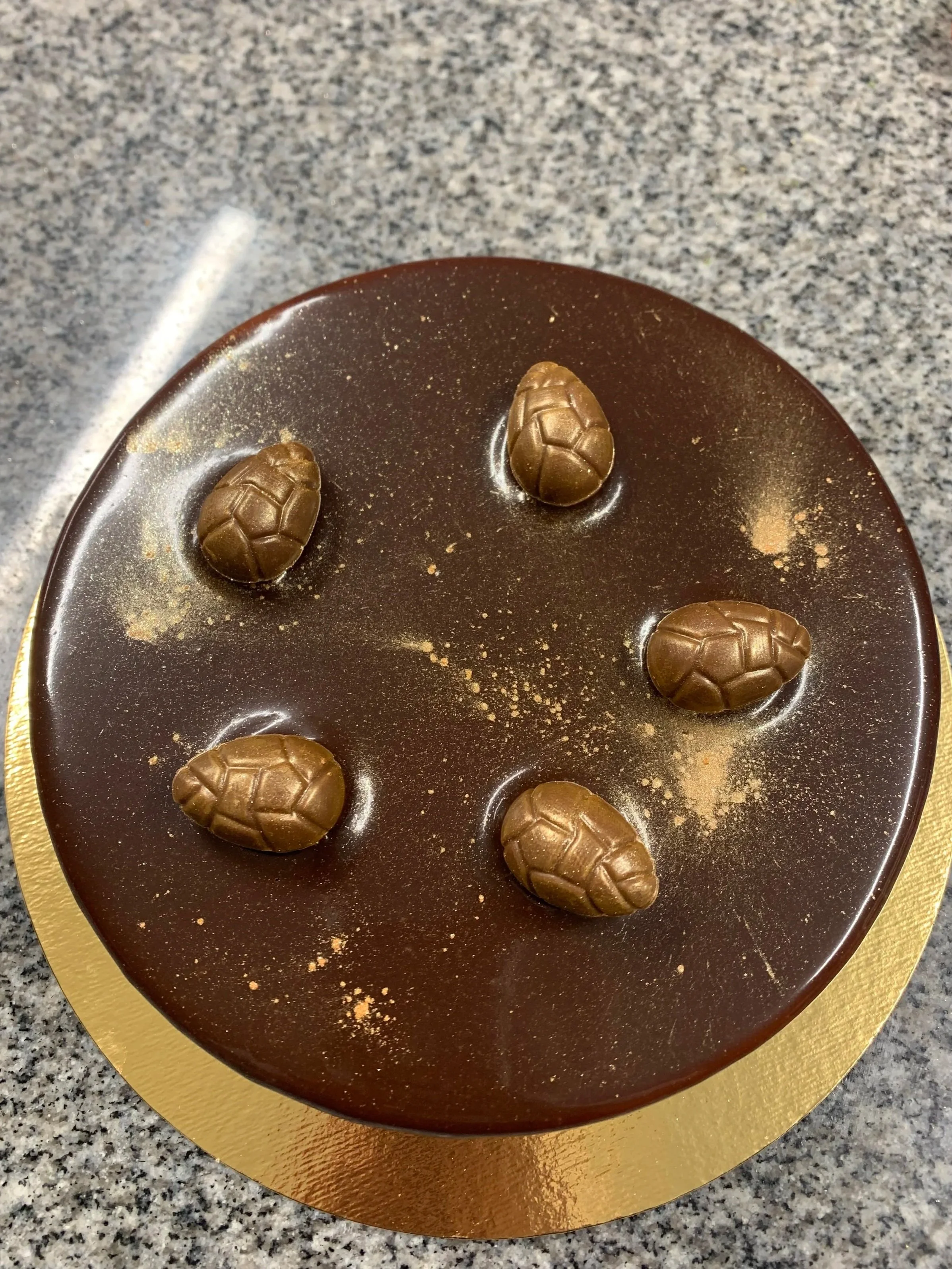 Gâteau au chocolat rond avec cinq petits œufs en chocolat à la surface, posé sur un plateau doré, sur une surface en granit.