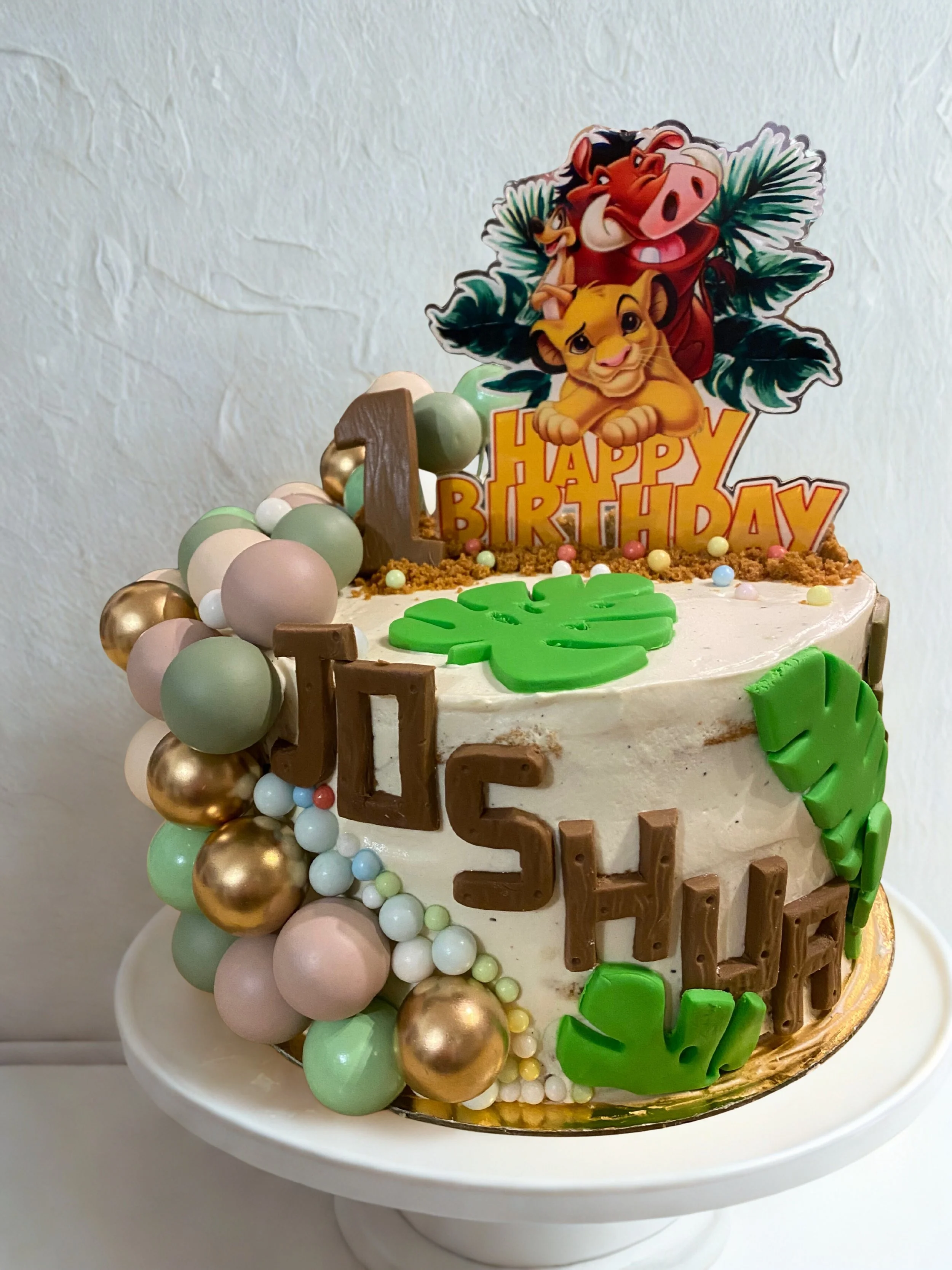 Gâteau d'anniversaire avec décorations de personnages du Roi Lion, incluant des ballons de différentes tailles et couleurs, et un message de fête en chocolat. Sur le dessus, une figurine en papier avec le texte 'Happy Birthday'.