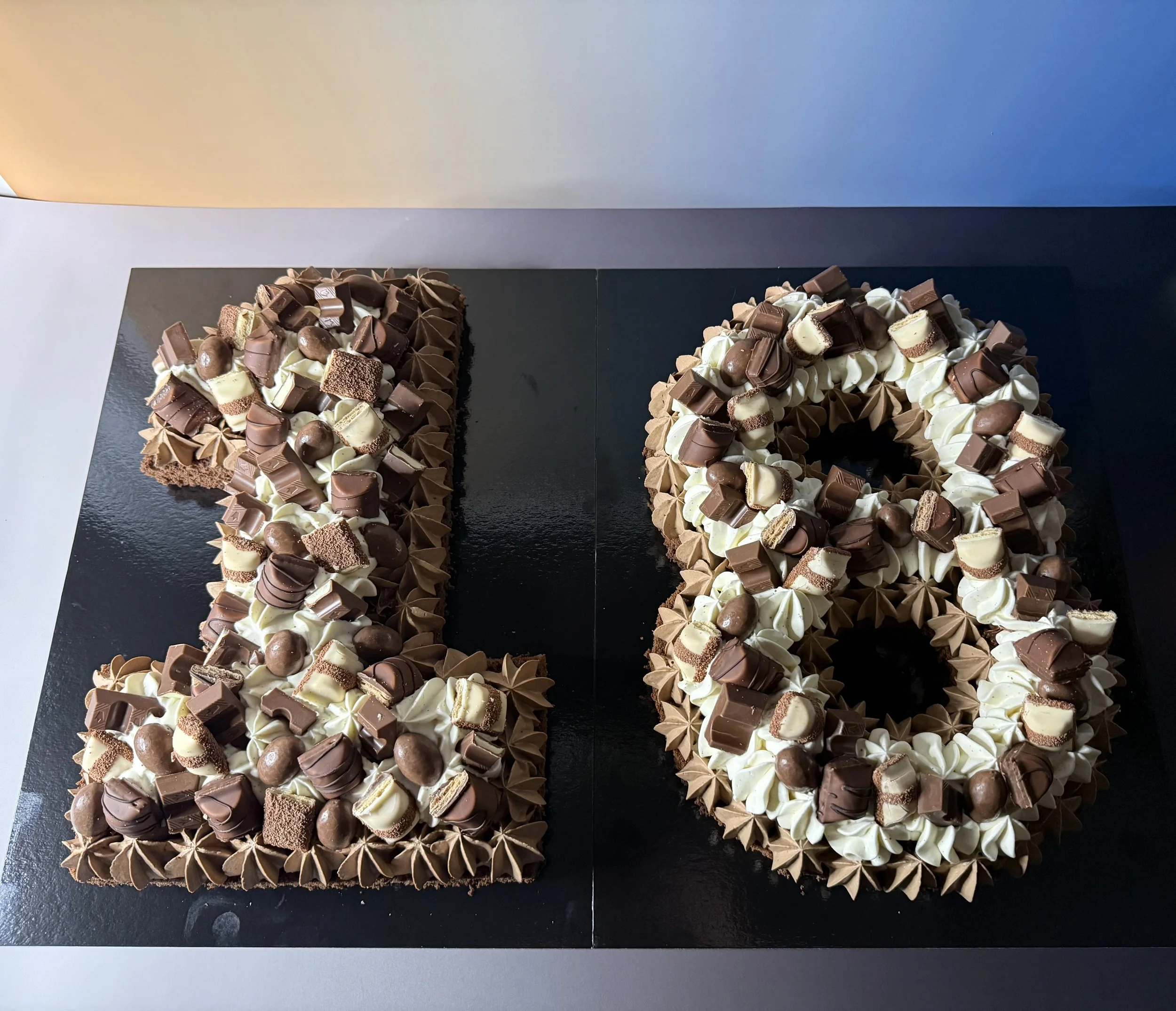 Gâteau en forme de chiffres '18', décoré avec divers chocolats et crèmes, sur une surface noire.