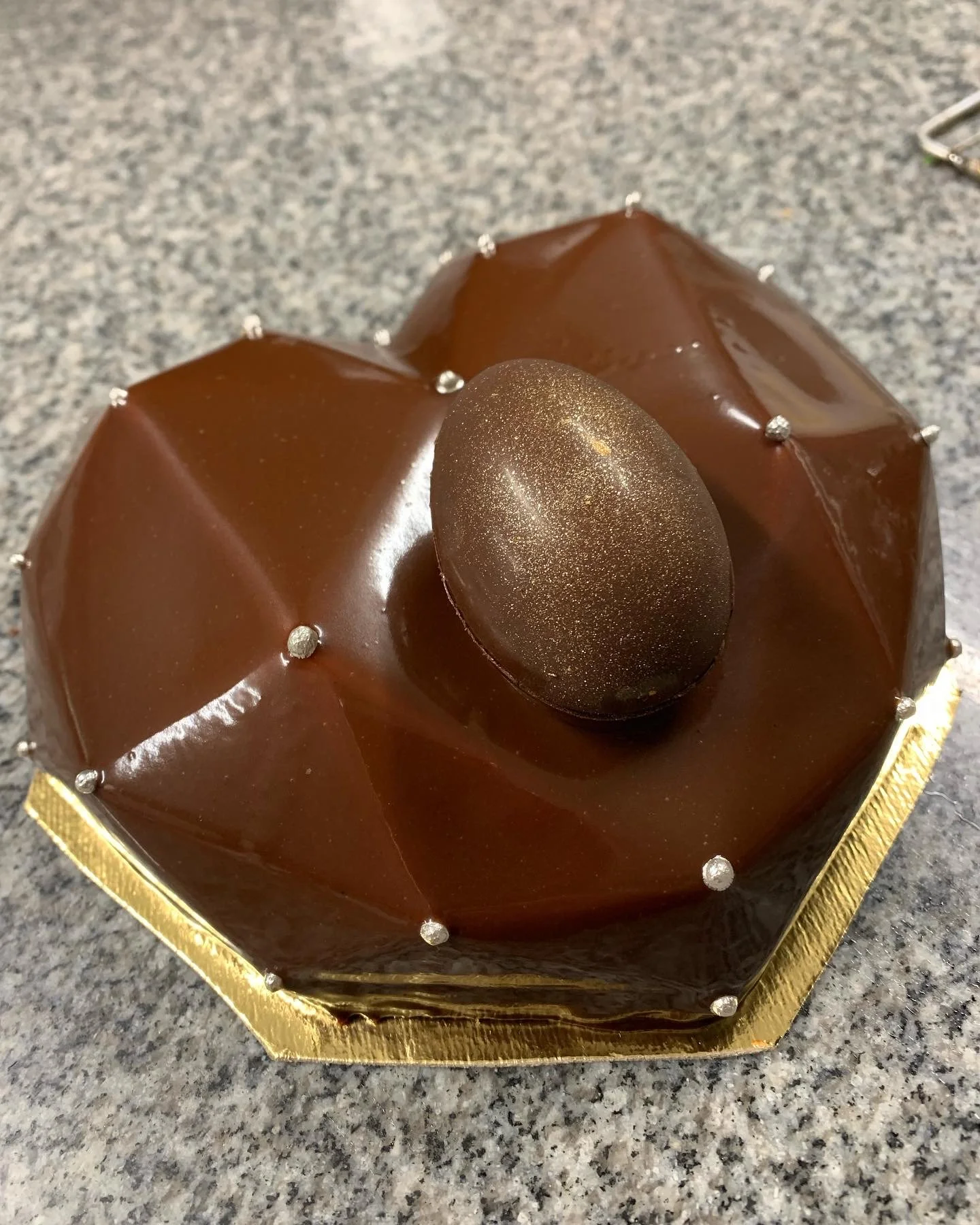 Gâteau au chocolat avec un œuf de Pâques en chocolat au centre, décoré de petites perles argentées, placé sur une surface en pierre.