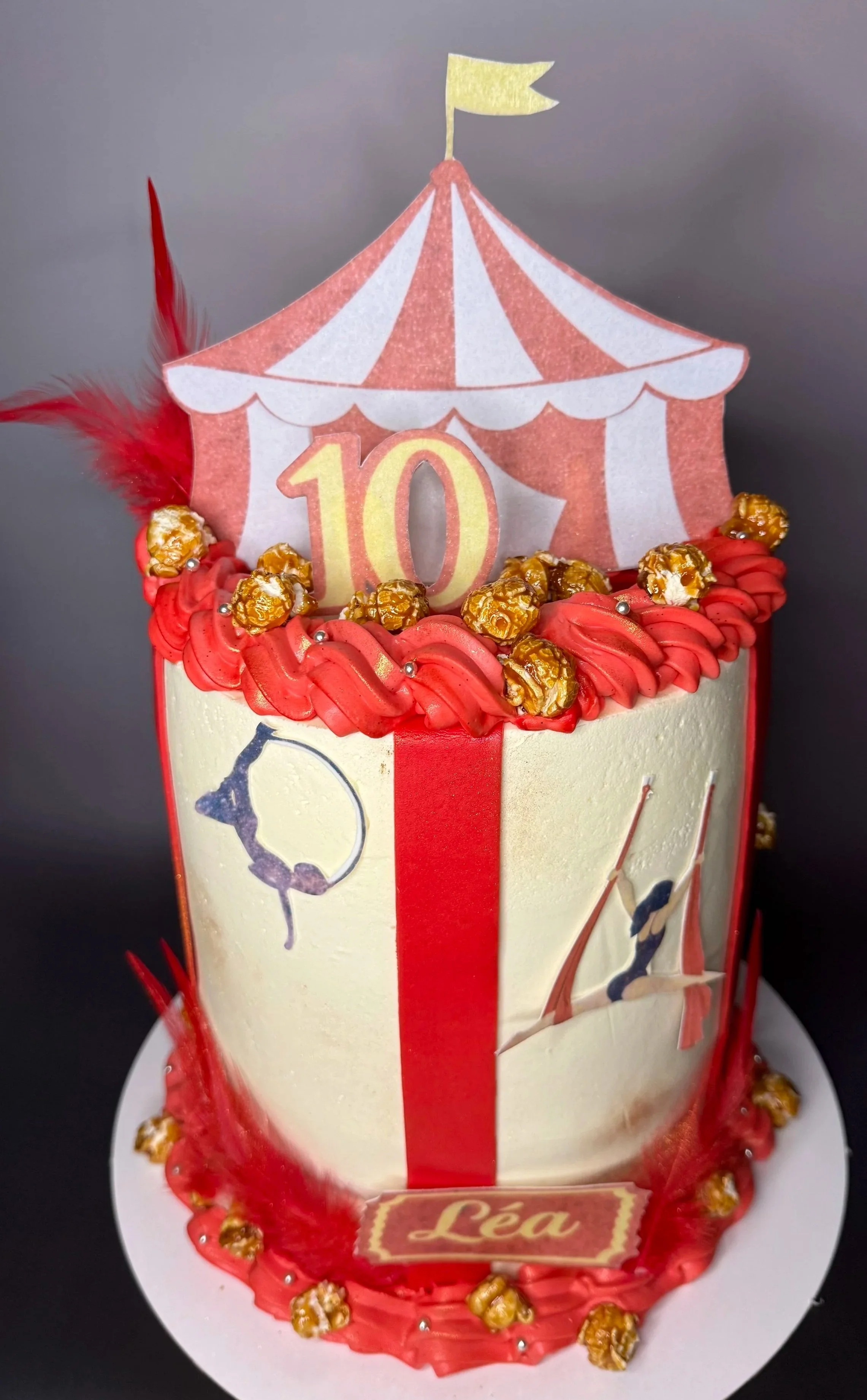 Gâteau anniversaire sur le thème du cirque, décoré avec une tente de cirque en papier, un chiffre 10 en relief, des popcorns en décoration, des détails en rouge et blanc, et une plaque avec le nom 'Léa'.