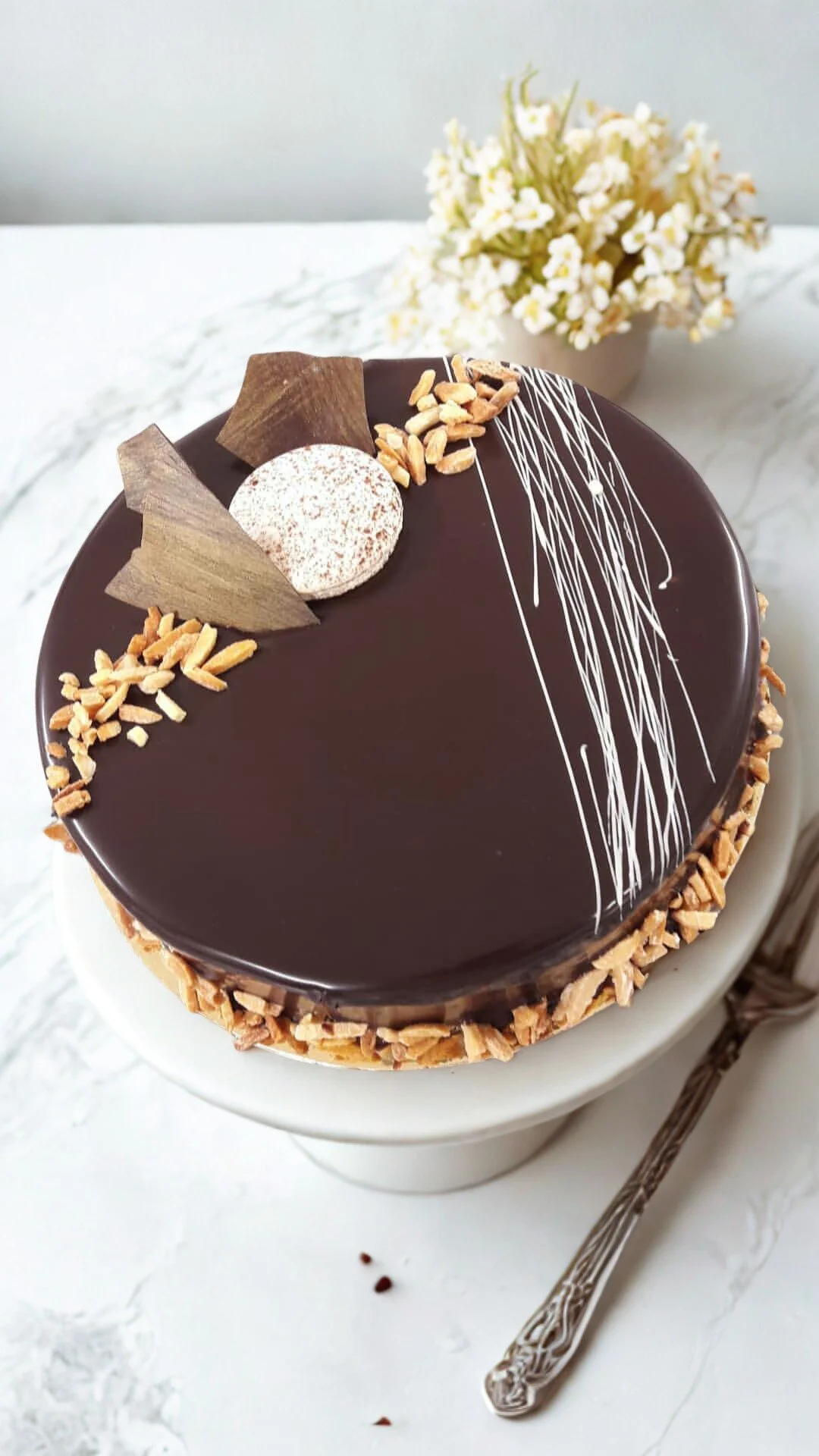 Gâteau au chocolat décoré avec des morceaux de chocolat, de la poudre de cacao, des éclats de caramel et des décorations en chocolat blanc, posé sur une table blanche avec une fourchette en argent à côté et un vase avec des fleurs blanches en arrière