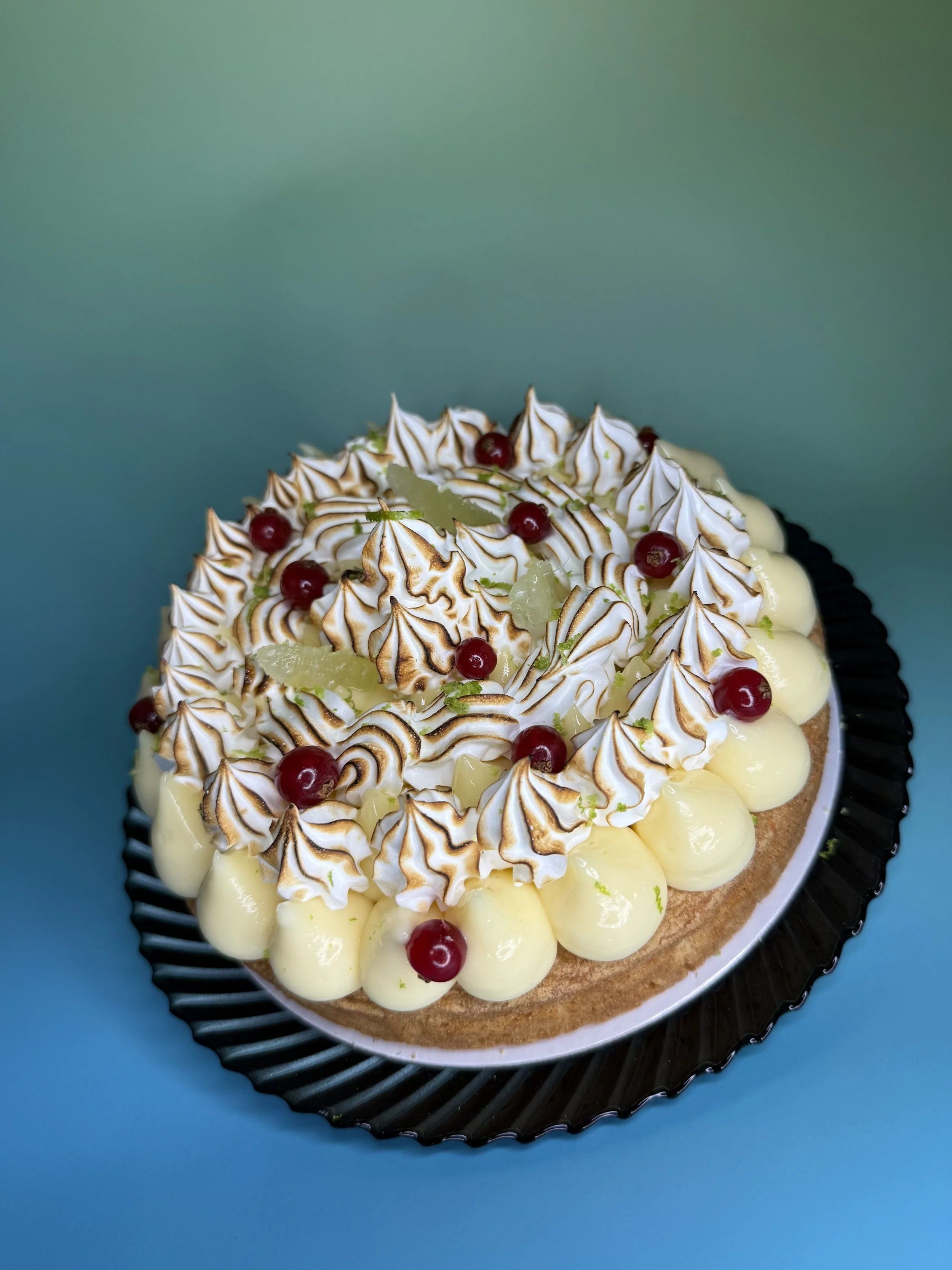 Gâteau décoré avec des meringues grillées, des groseilles, des morceaux de citron vert confit, et de la crème à la vanille, sur une assiette noire sur fond bleu.