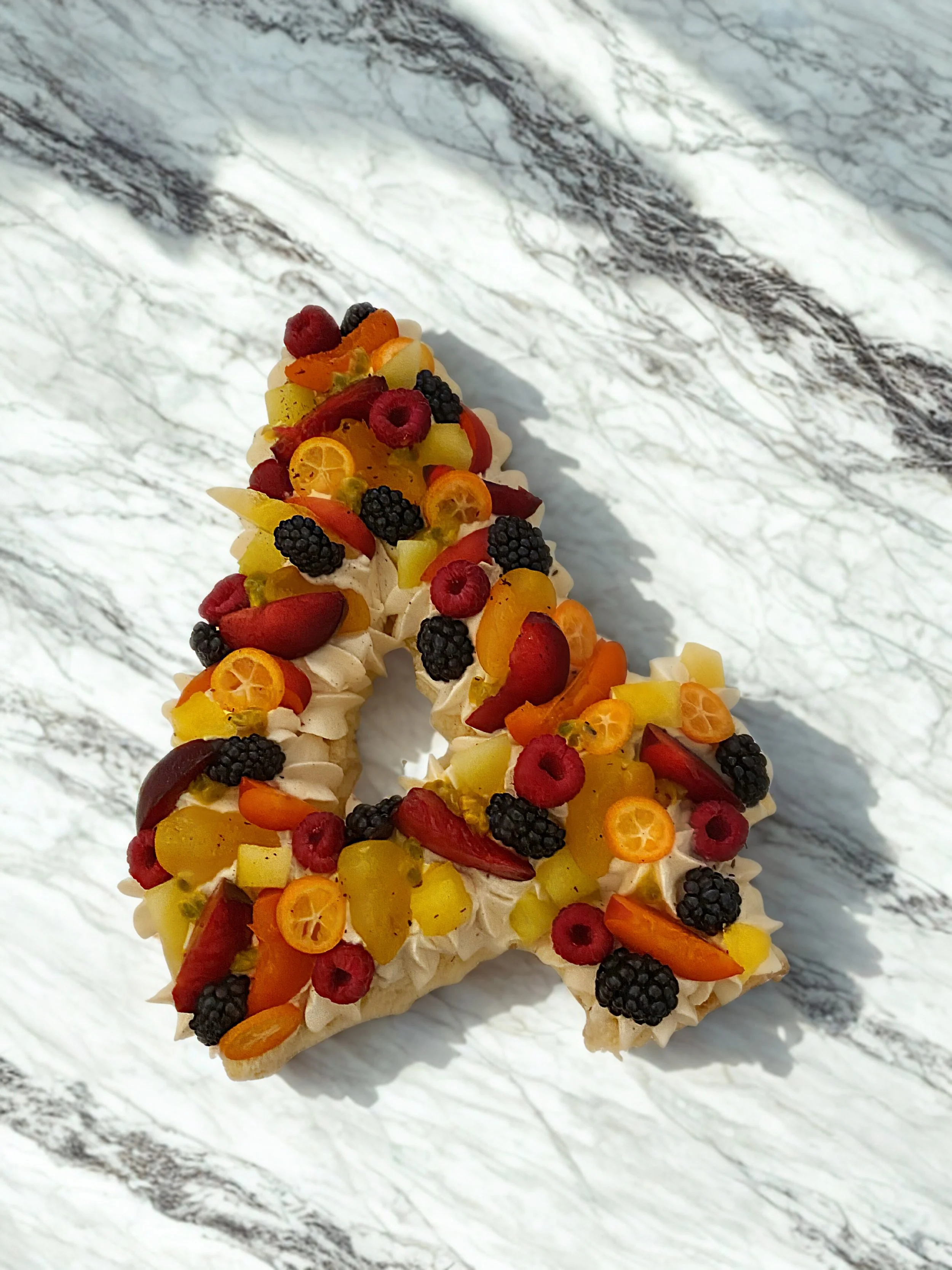 Gâteau en forme de cœur décoré avec des fruits frais comme des fraises, des framboises, des mûres, des tranches de kumquat, des morceaux d'ananas et d'autres fruits, sur une surface blanche avec un motif marbré.