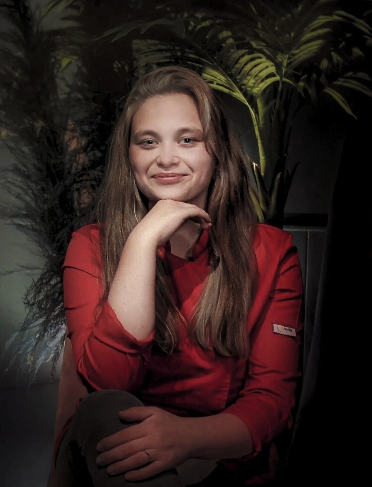 Jeune fille avec des cheveux longs portant une chemise rouge, assise devant des plantes vertes, souriant et regardant vers l'avant.