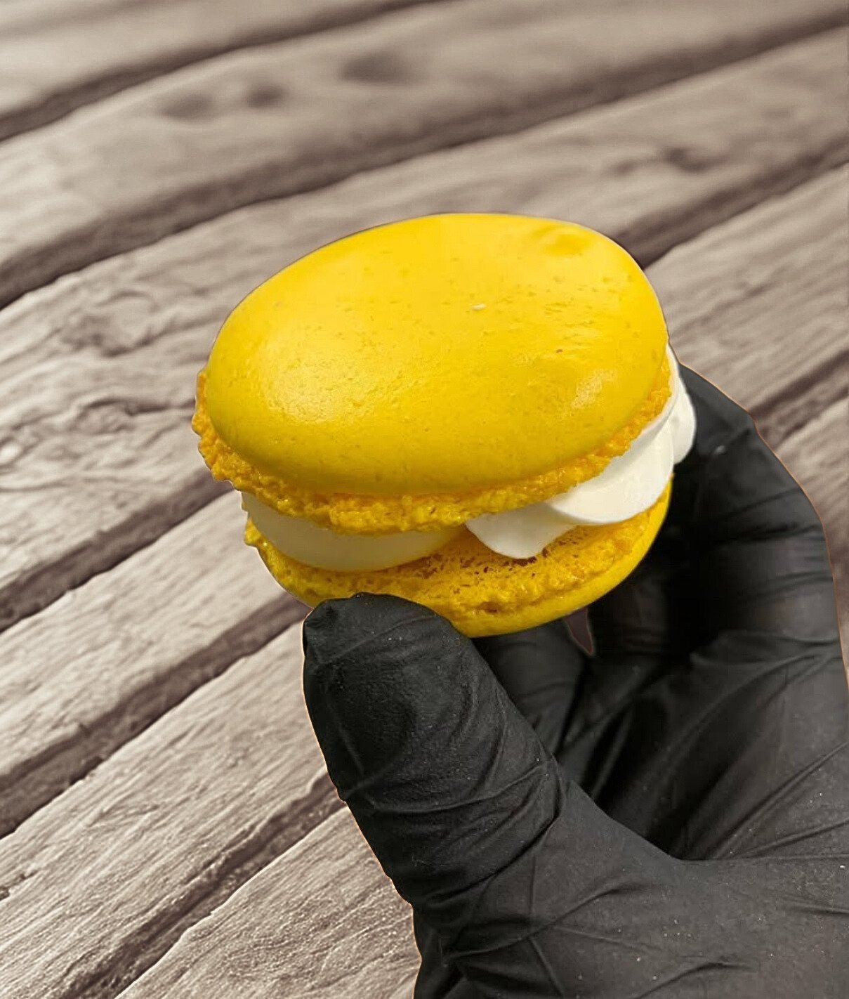 Macaron jaune avec crème blanche à l'intérieur, tenu par une main portant un gant noir, posé sur une surface en bois.